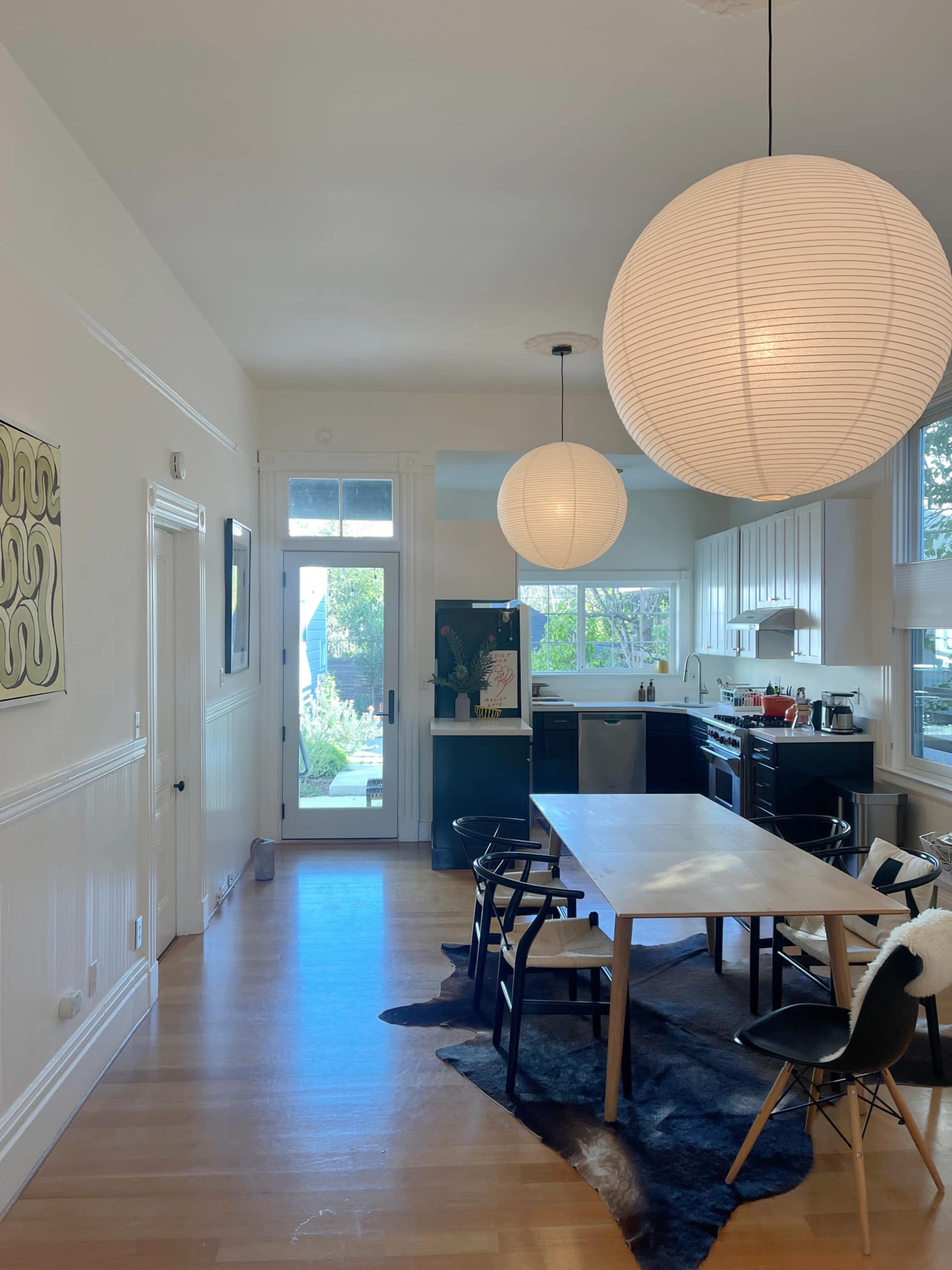 A well-lit kitchen features a dining table with black chairs, two large pendant lights overhead, and an open view of the outdoors through a glass door.