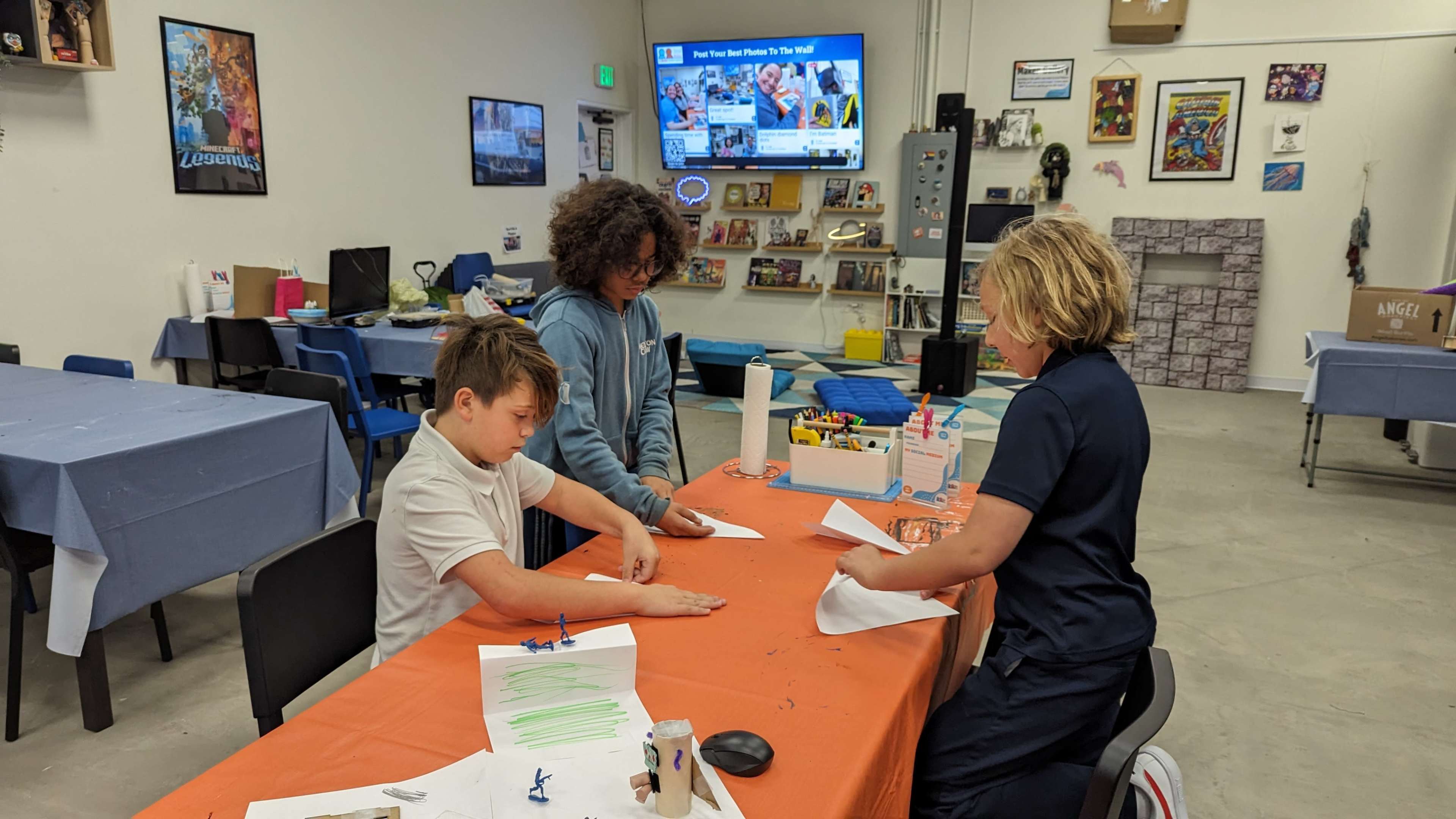 Three children engage in a crafting activity at a table in a room with a television displaying a game.