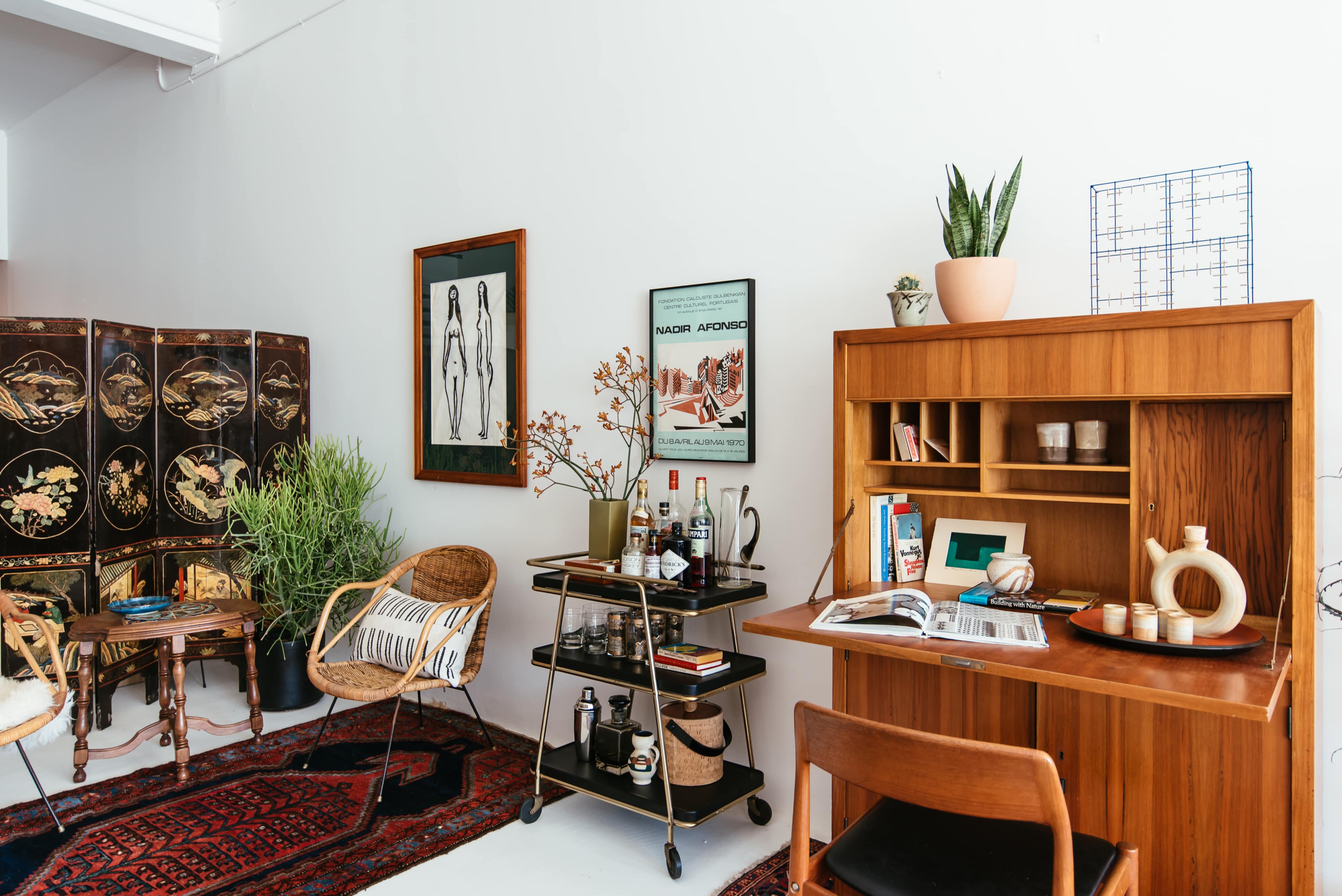 A stylish interior living space featuring a wooden desk, a bar cart, a variety of plants, and decorative wall art.