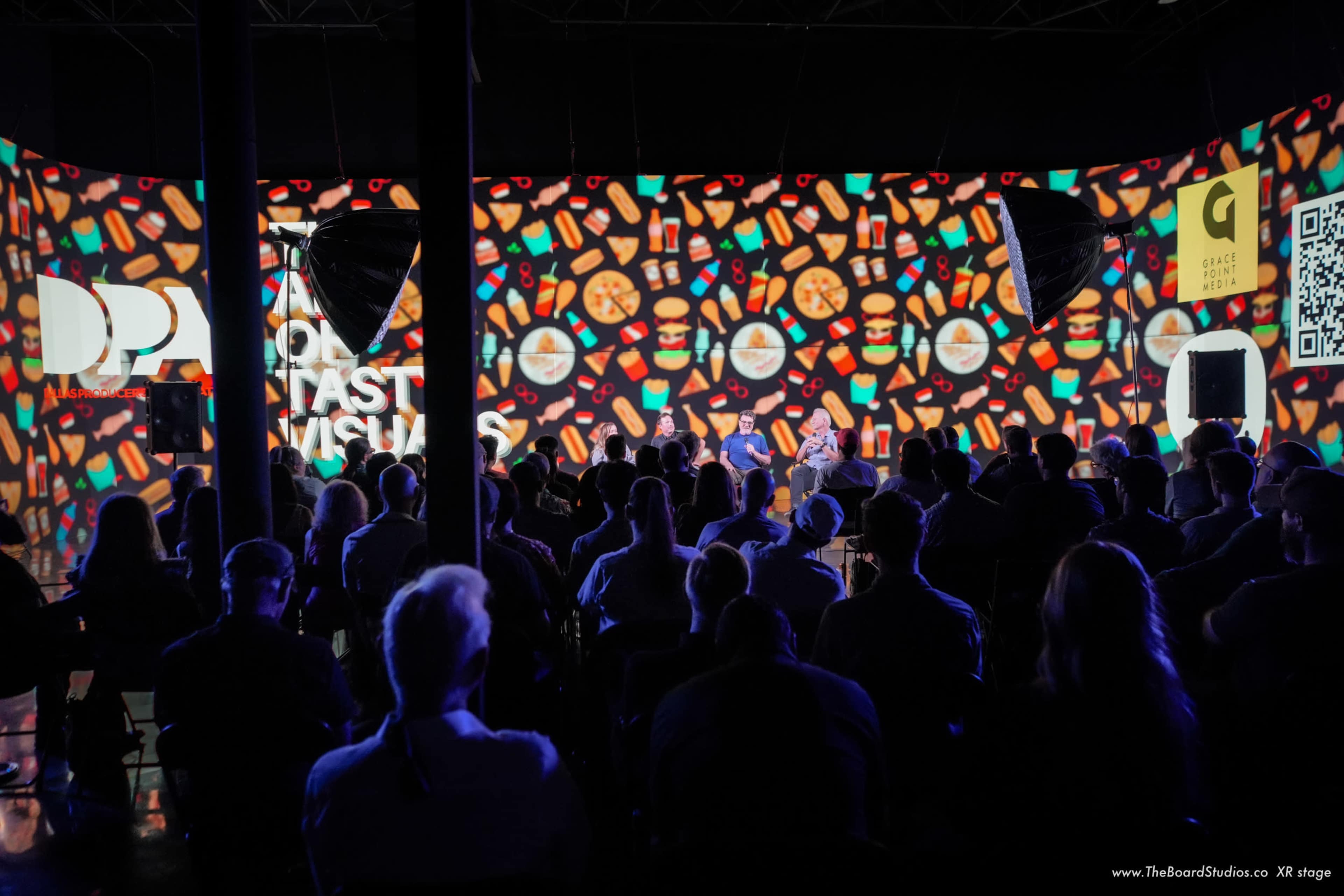 A crowd of attendees sits in front of a colorful, patterned backdrop during a presentation at an event.