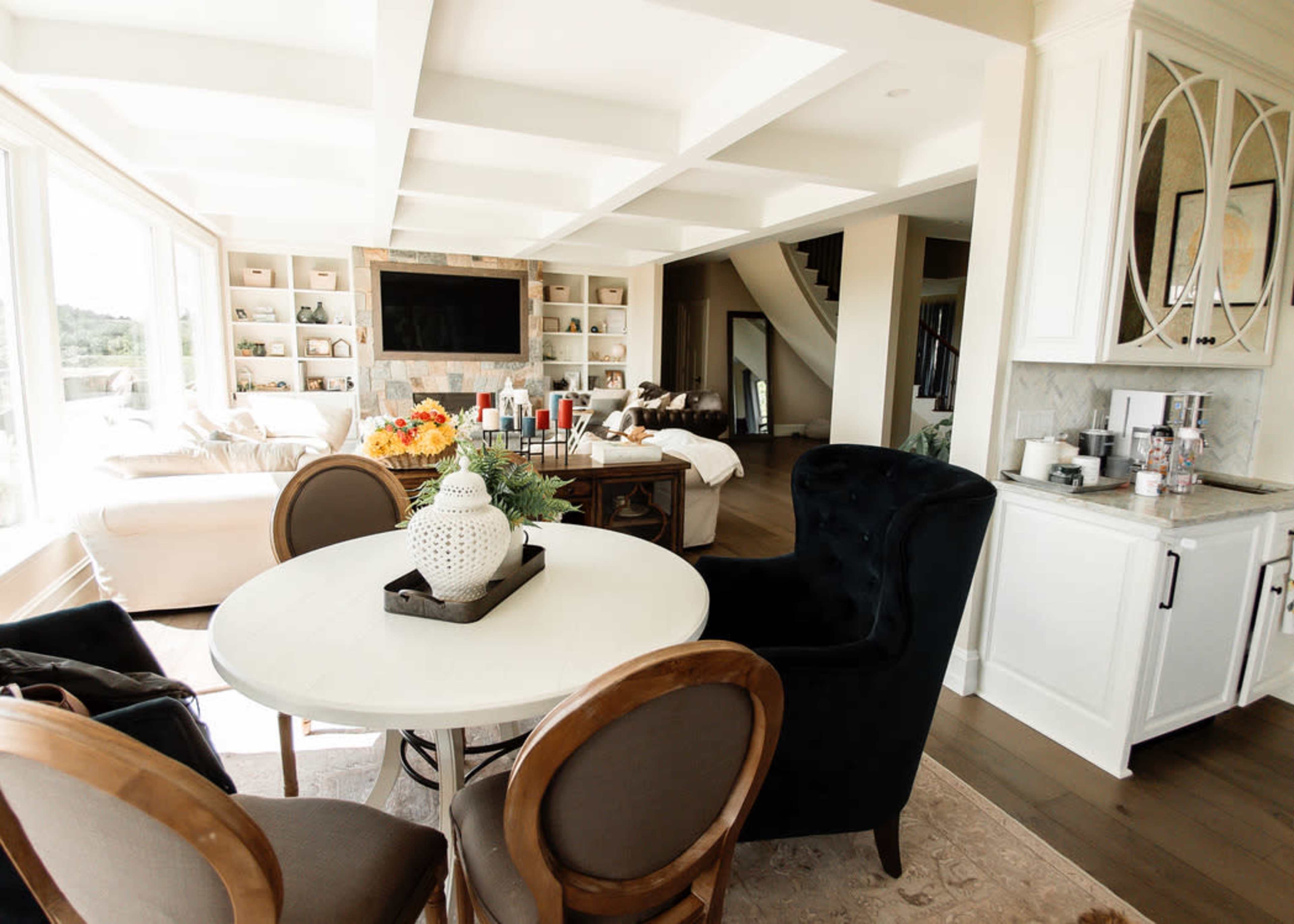 A modern dining area features a round table surrounded by chairs, with a light-filled open space leading to a cozy living area in the background.