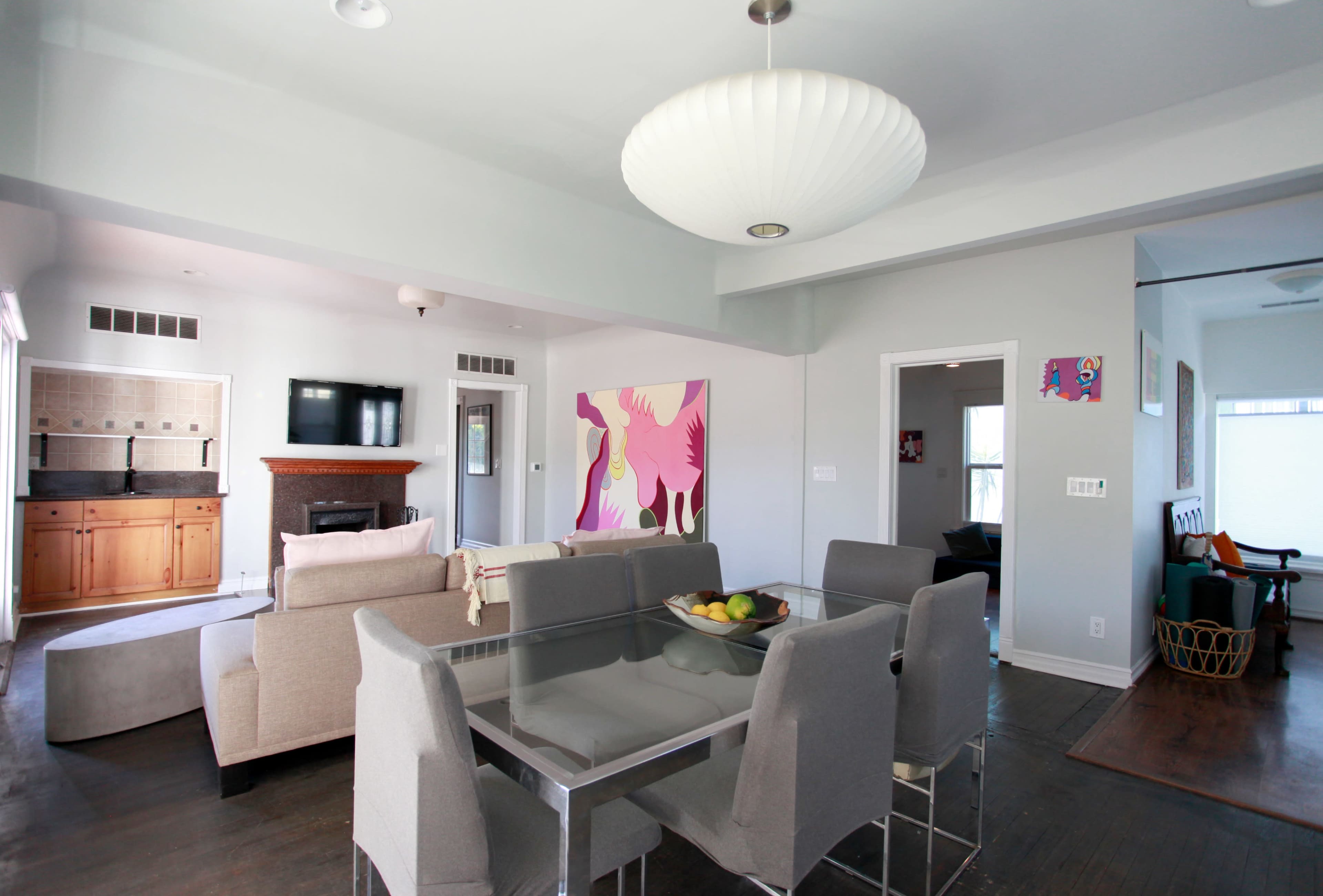 The image shows a modern, open-concept living space featuring a dining table with gray chairs, a sofa area, and artwork on the walls.