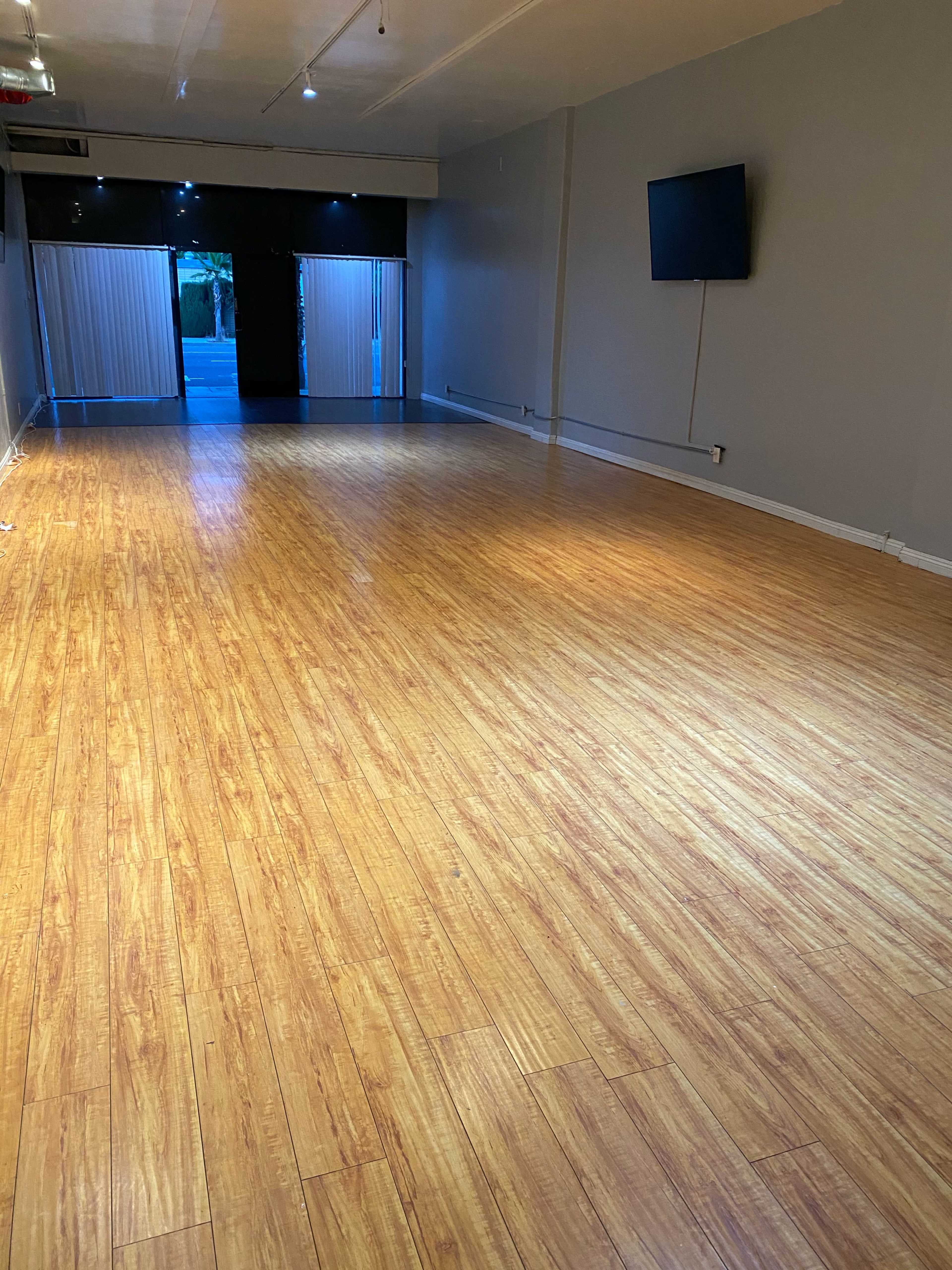The image shows a spacious room with hardwood flooring, minimal furnishings, and a large window providing natural light.