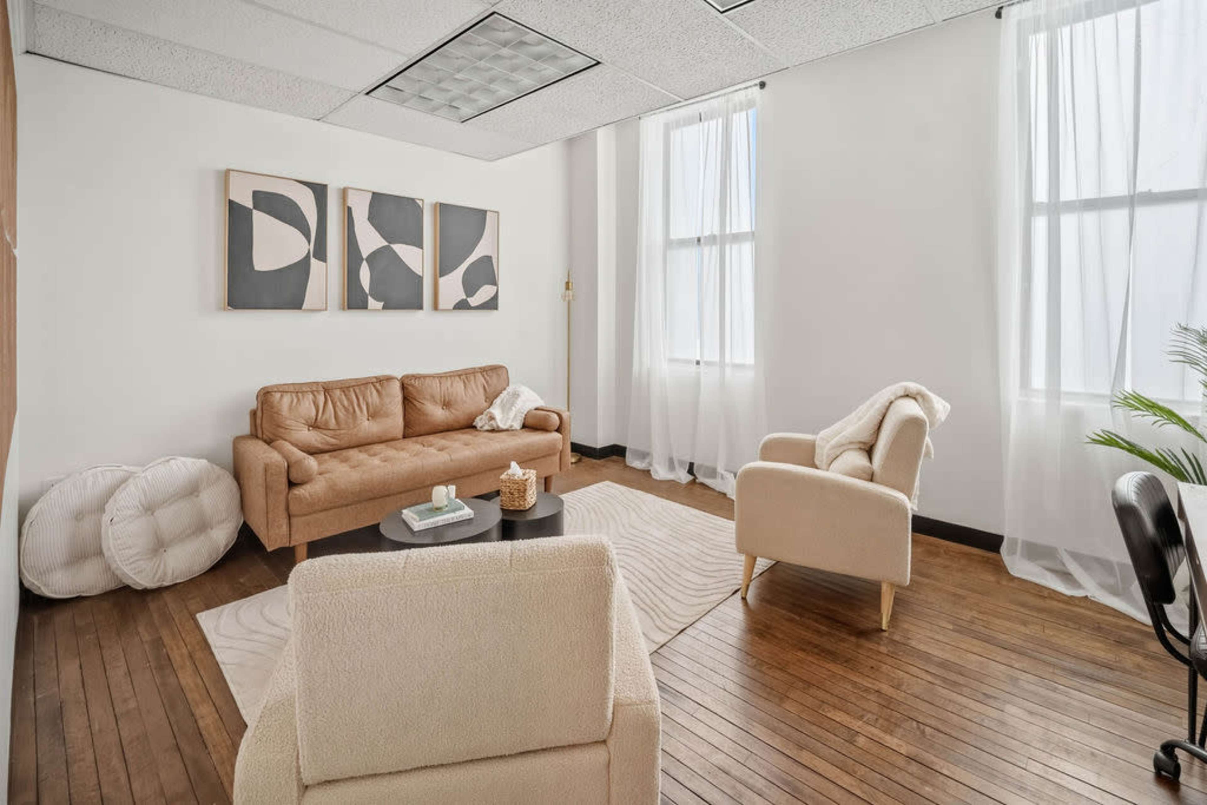 The image shows a bright, minimalistic office space with a tan leather sofa, a cream-colored armchair, and a small black coffee table, decorated with abstract wall art and natural light filtering through sheer curtains.