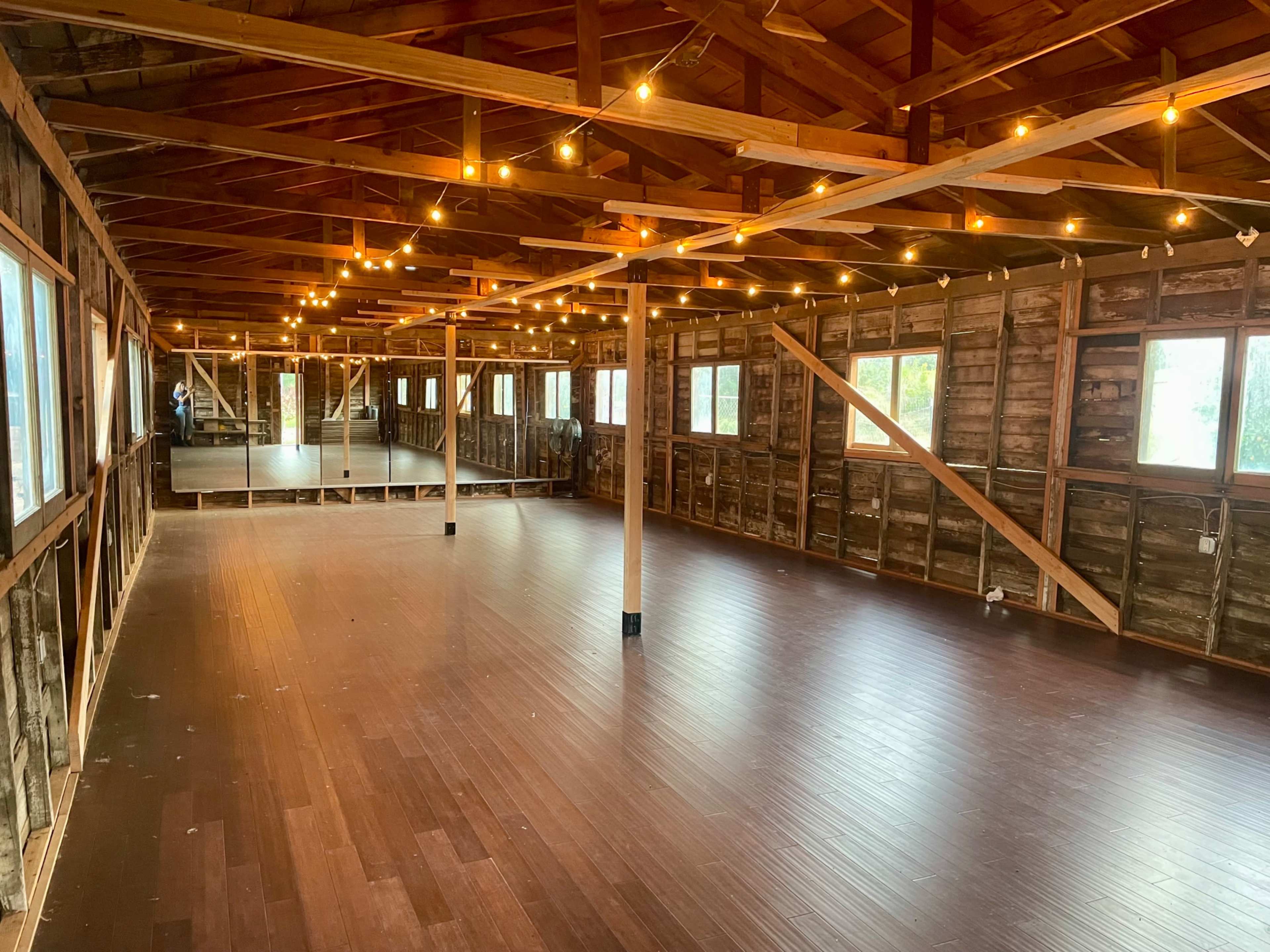 The image shows a spacious, rustic interior with wooden beams and flooring, illuminated by string lights and featuring several windows.