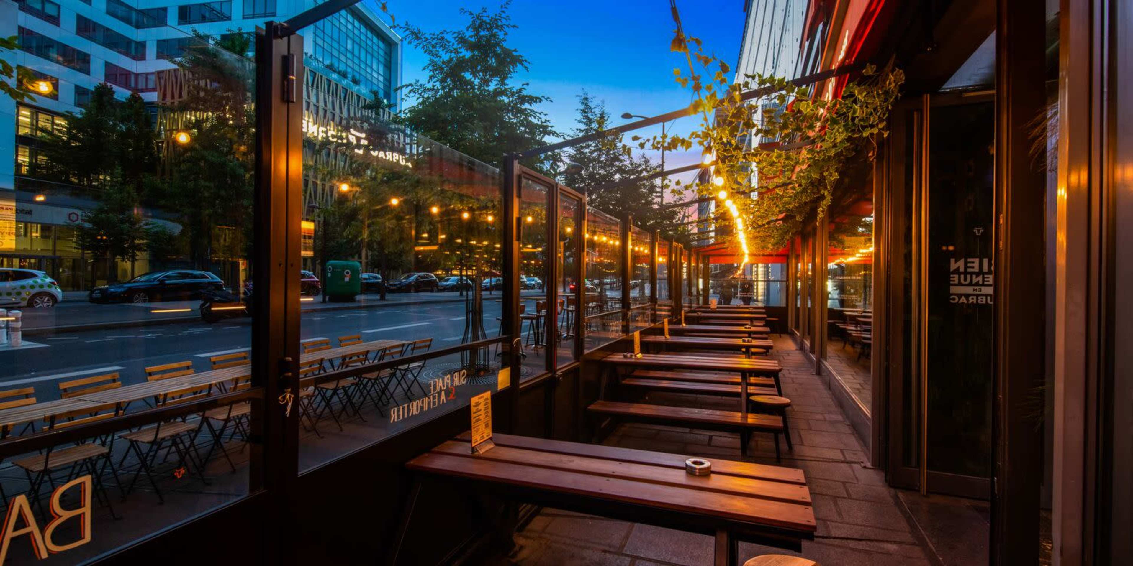 An outdoor café seating area with wooden benches lines a city street, illuminated by string lights at dusk.