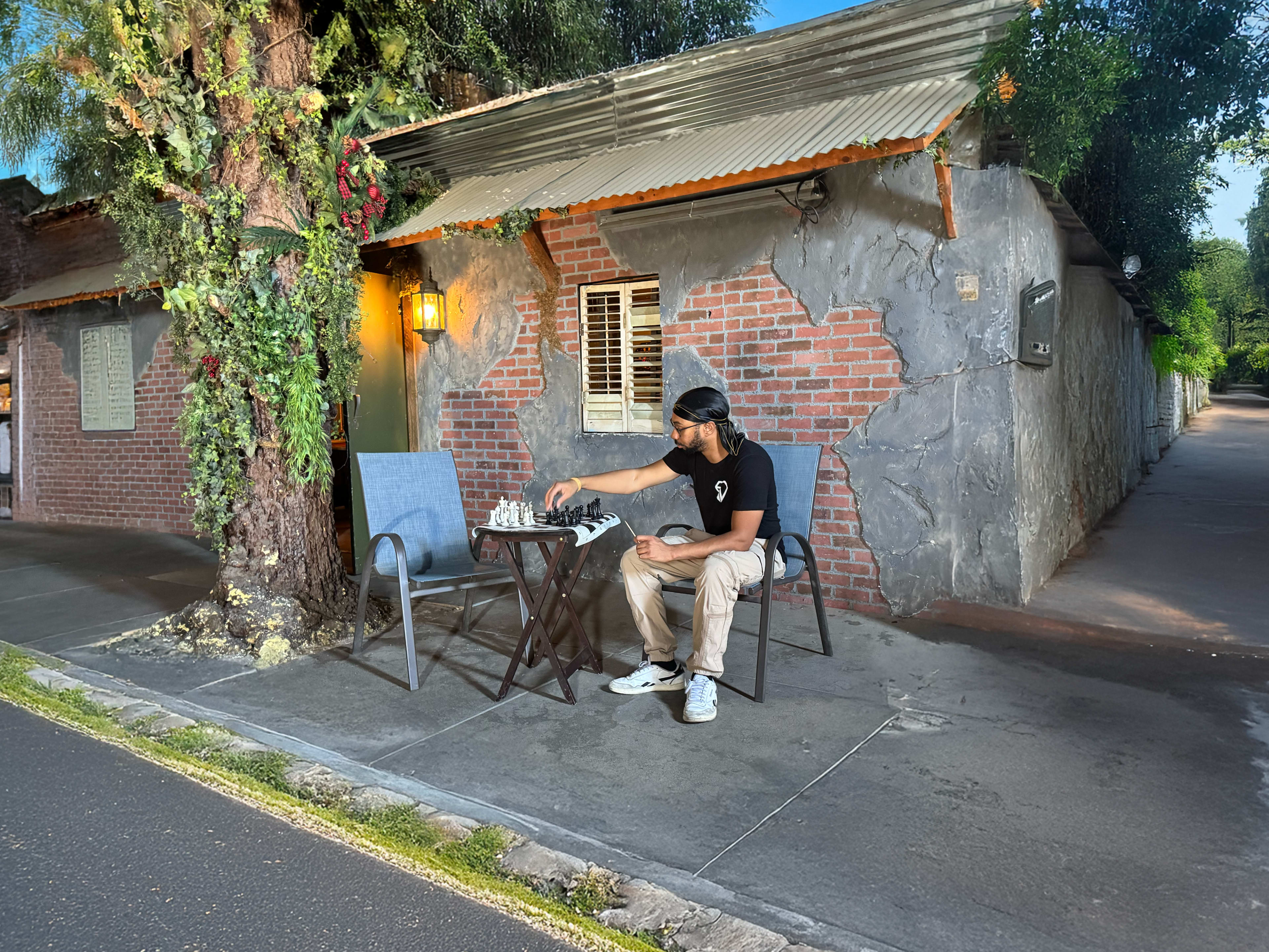 A person sits at a table outside a rustic building, playing chess on a sunny day.