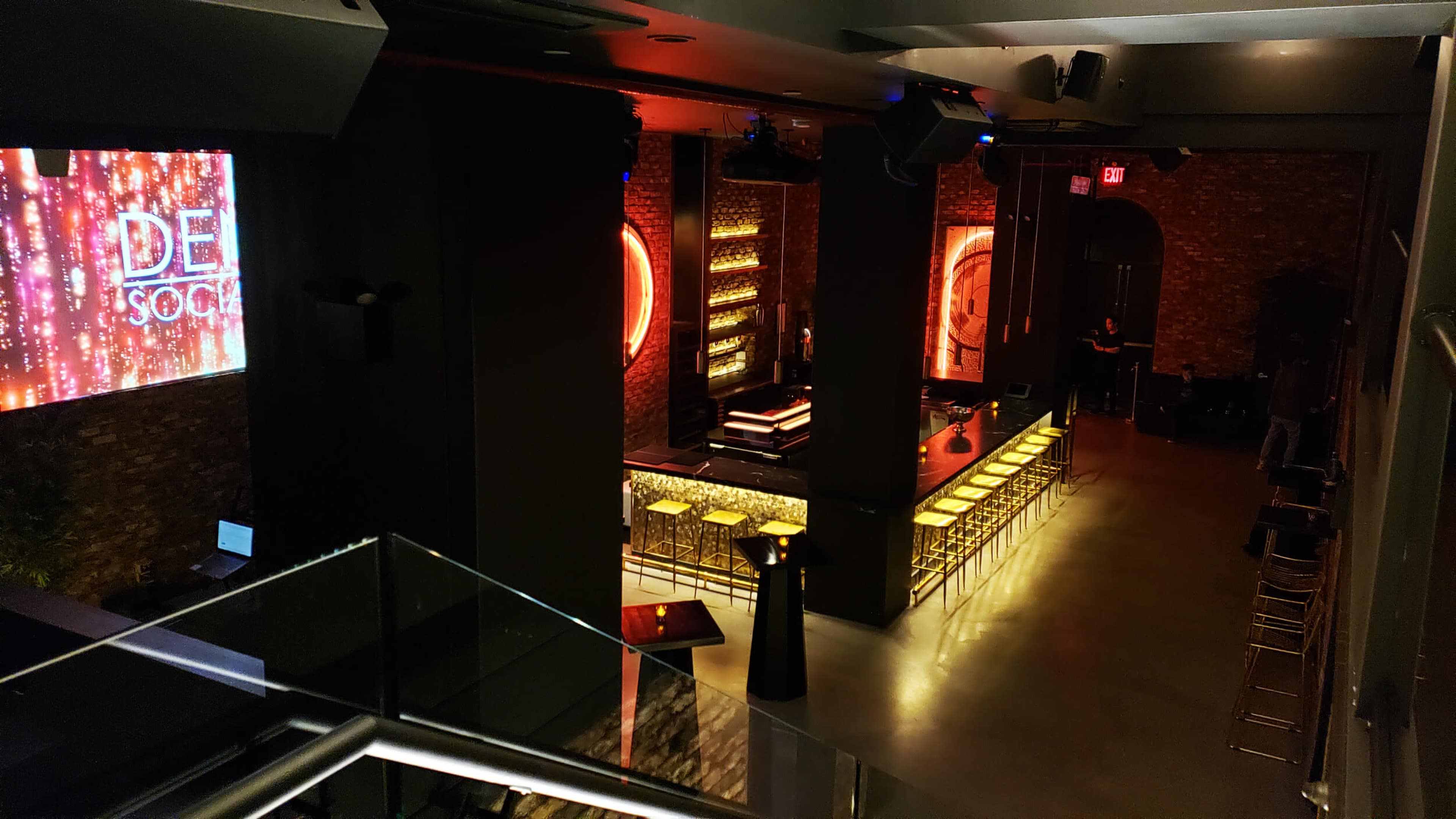 A dimly lit bar area with a neon-lit backdrop, featuring a long row of gold bar stools and a display screen on the wall.