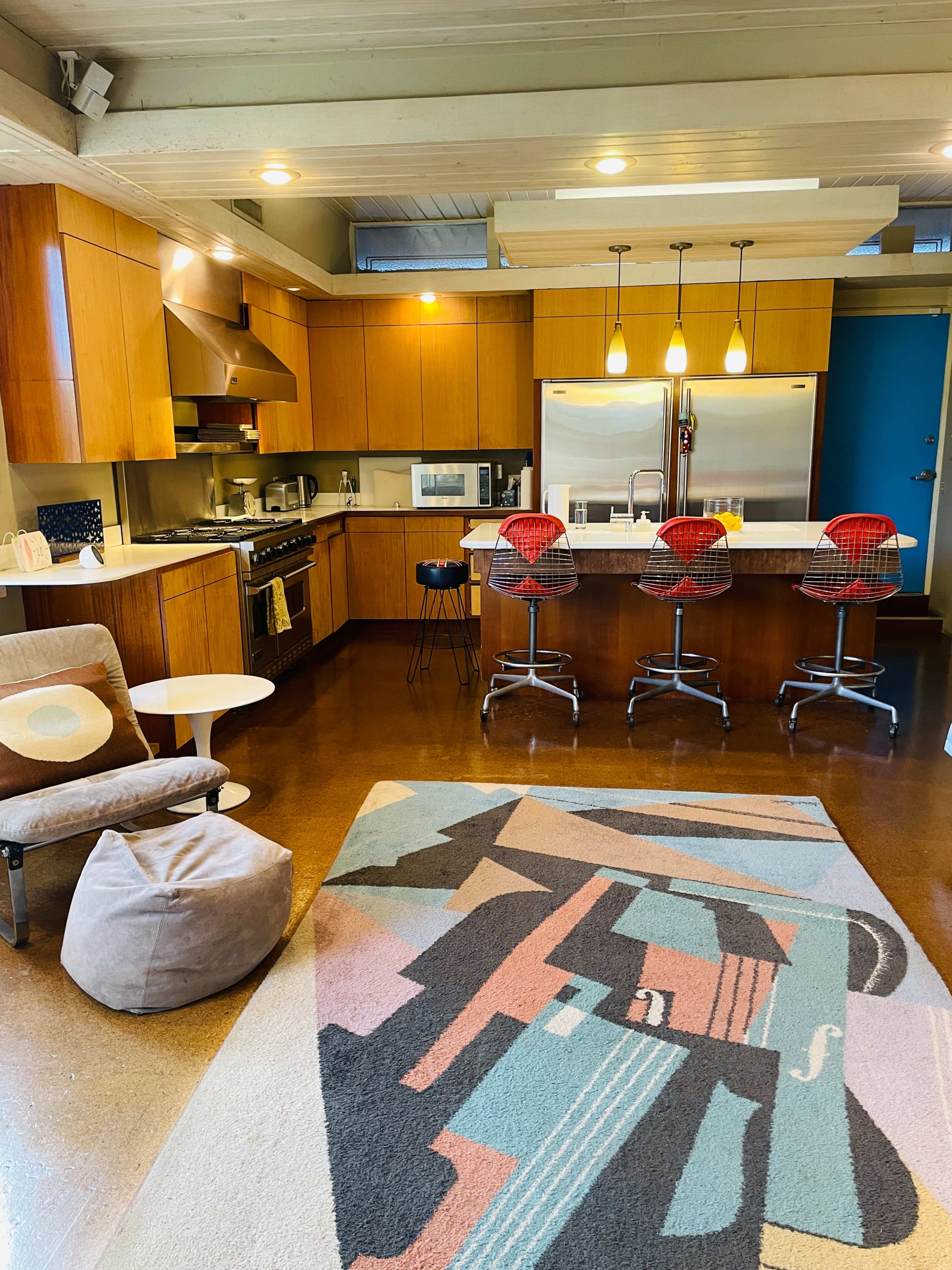 The image shows a modern kitchen with wooden cabinetry, three red bar stools, and a colorful area rug.