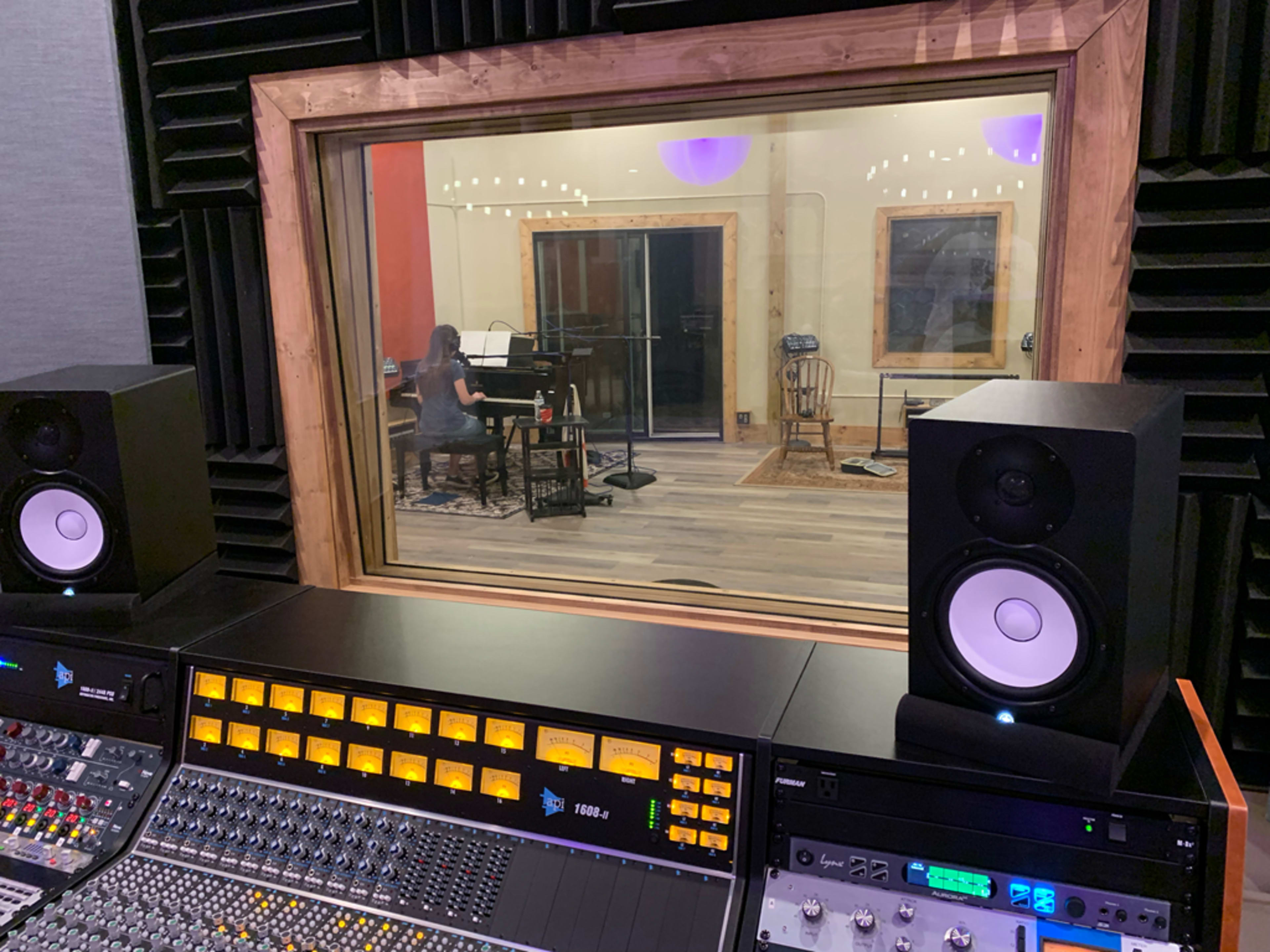 A music recording studio features a control panel in the foreground and a musician playing an instrument visible through a window in the background.