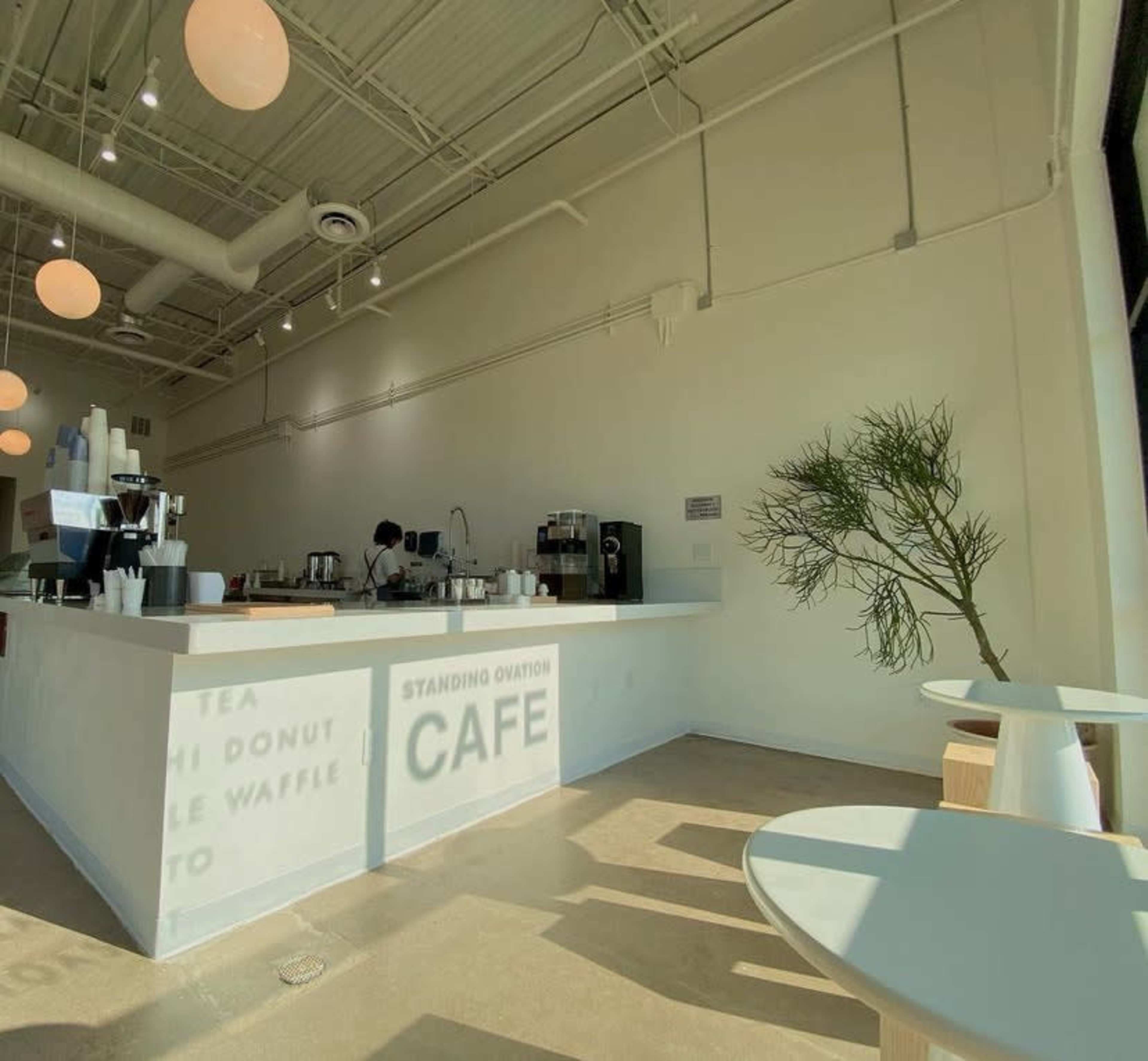 The image depicts a modern, minimalist café interior featuring a white service counter, light wooden furniture, and a barista preparing drinks in the background.
