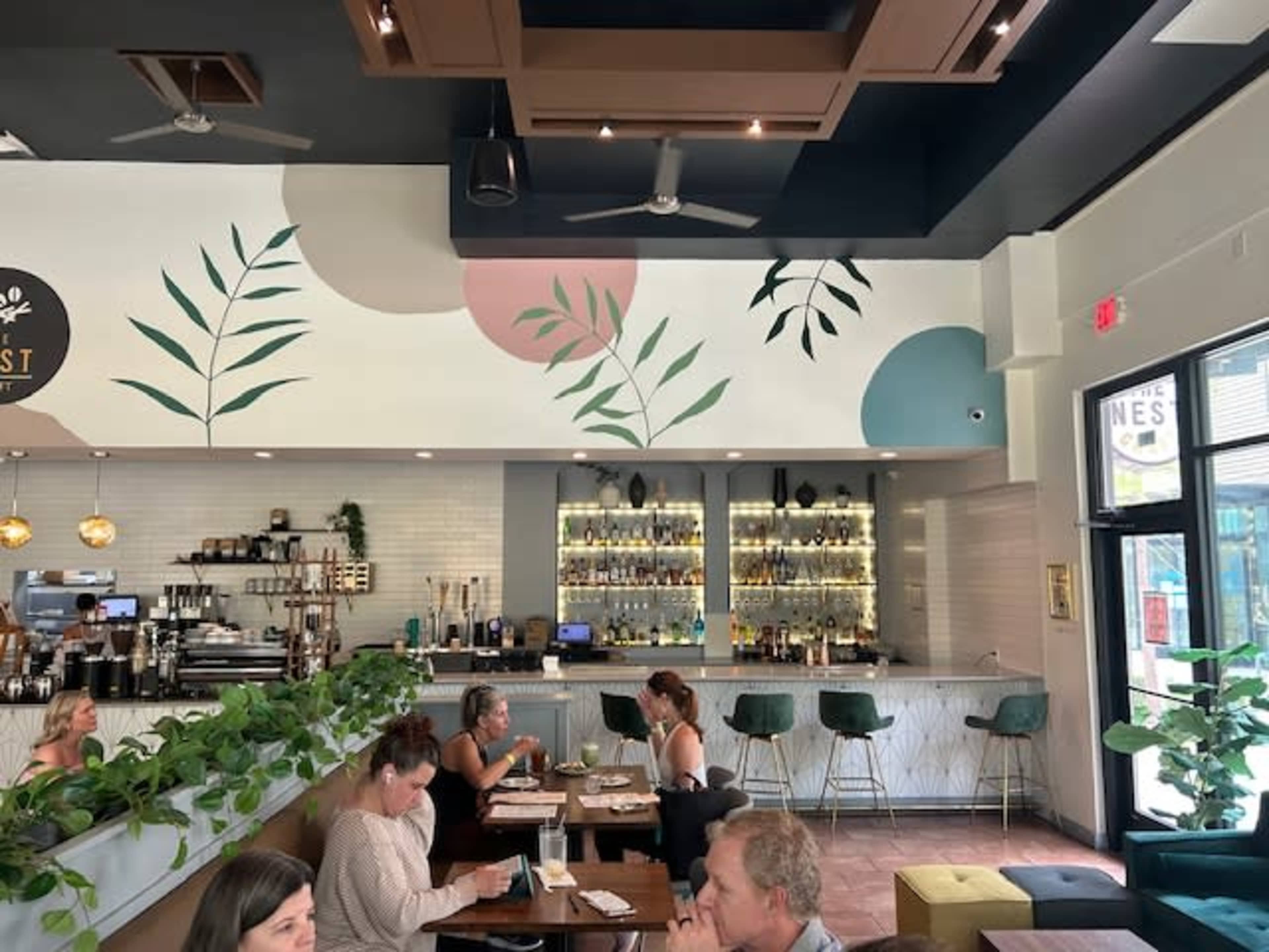 The interior of a modern café features a bar area with a variety of spirits on display and leafy wall decor, while patrons occupy tables and enjoy their meals.