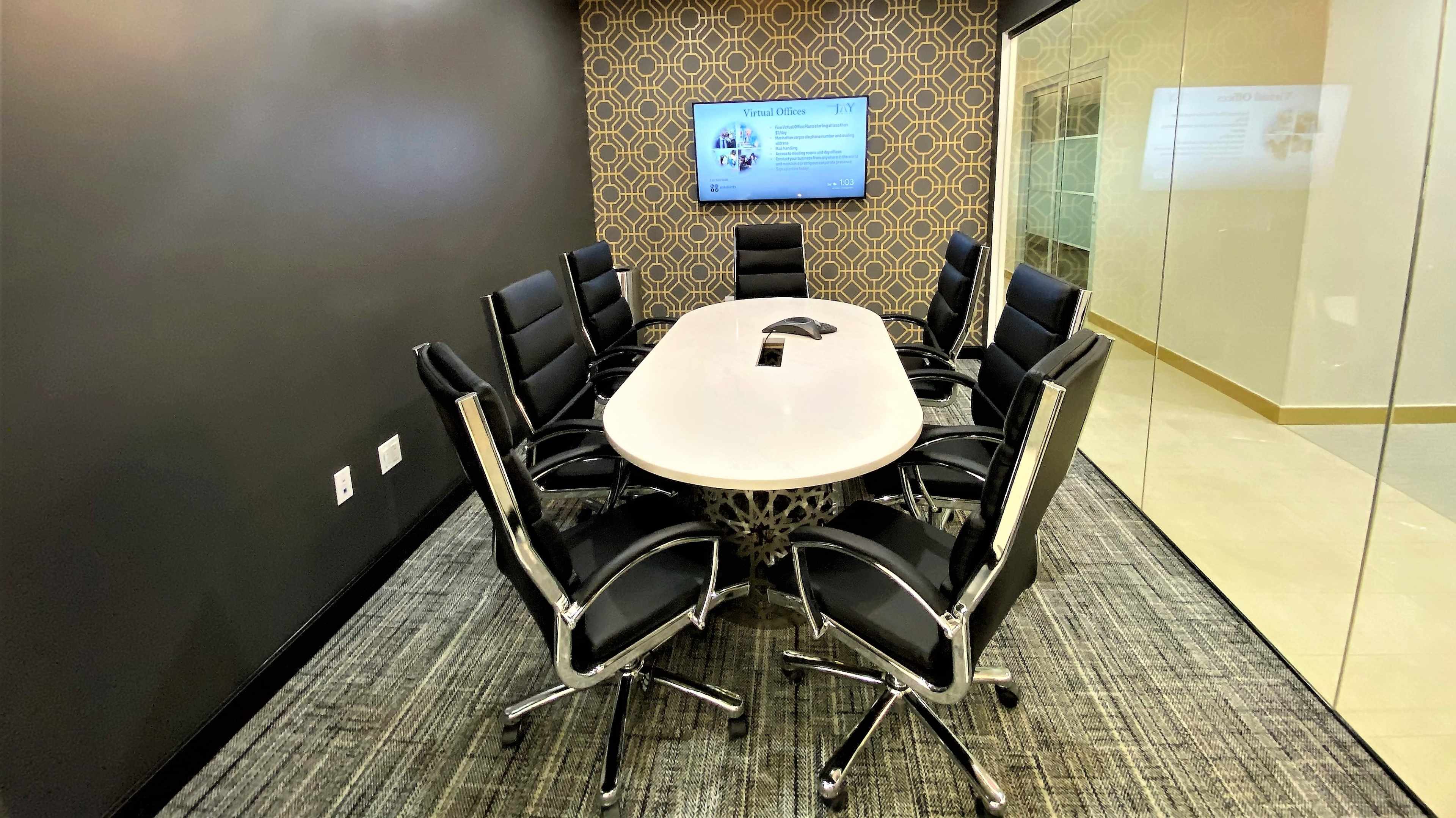 A modern conference room features a large oval table surrounded by black chairs and a wall-mounted screen.