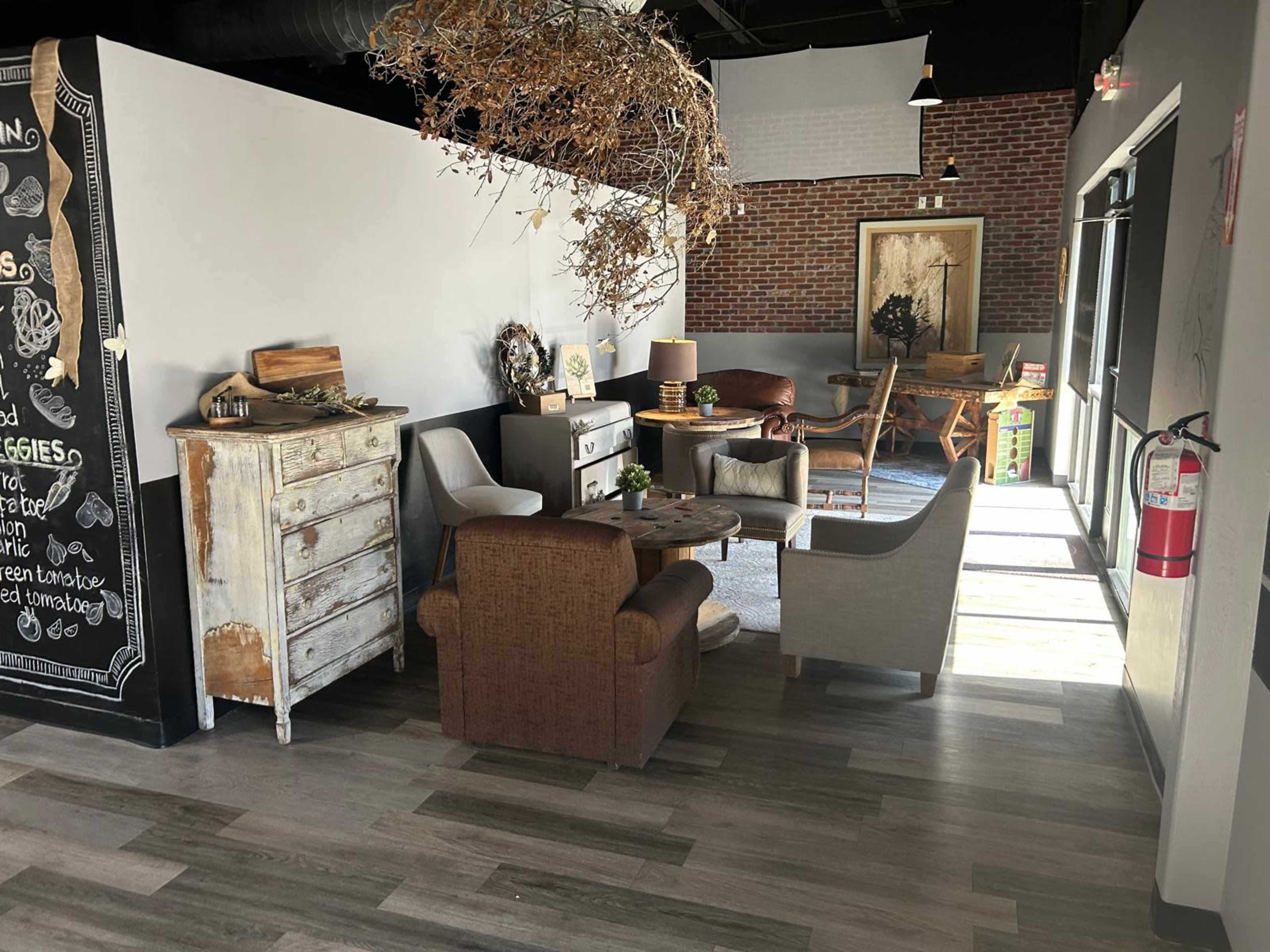 The image shows a cozy seating area with various chairs and a wooden table, surrounded by decorative elements such as a rustic dresser and a wall adorned with greenery.