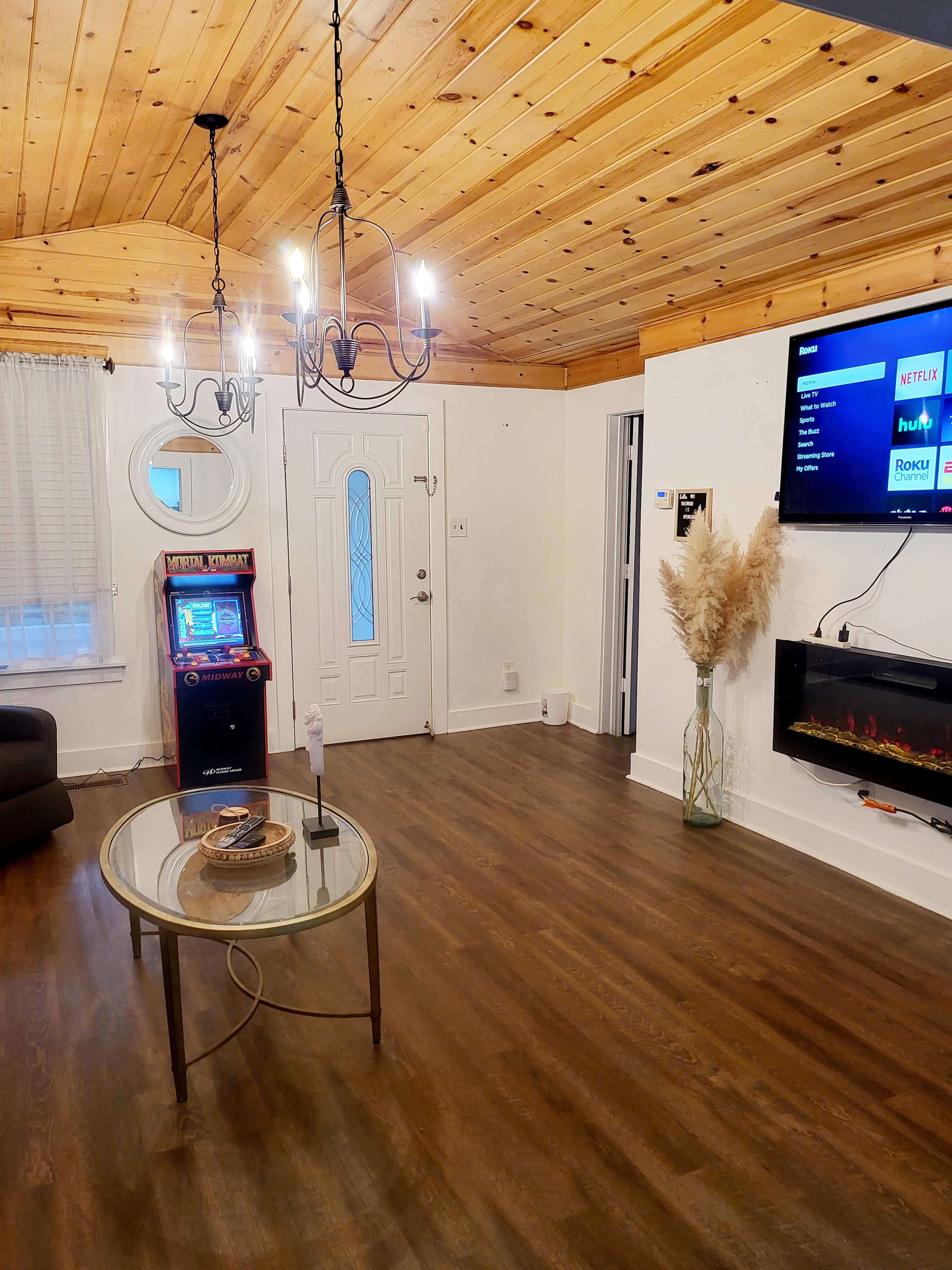 The interior of the room features a wood-paneled ceiling, a front door, an arcade game machine, a modern electric fireplace, and a round table with a decorative item on it.
