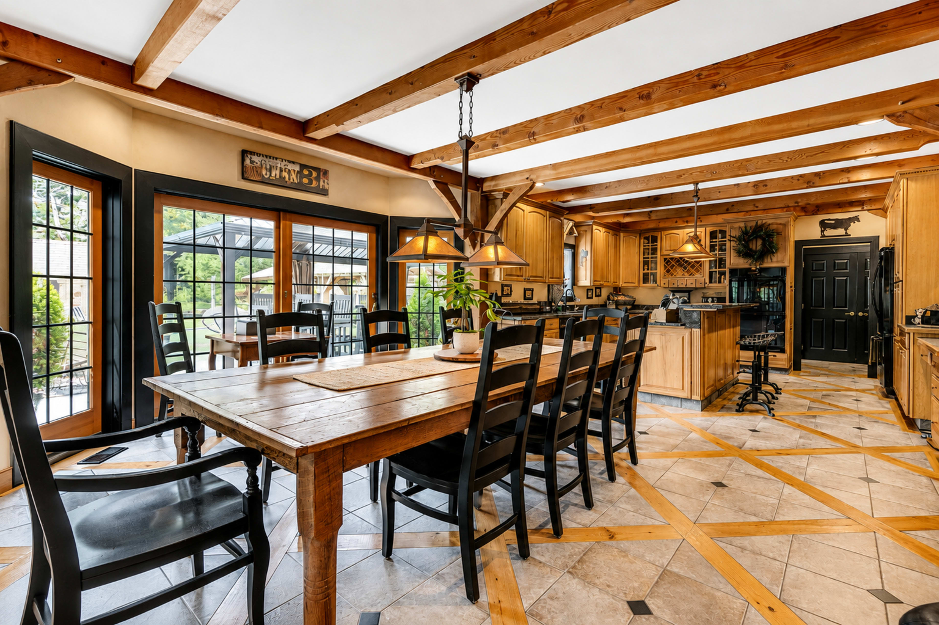 The image shows a spacious kitchen and dining area with wooden beams, a large table surrounded by black chairs, and multiple windows allowing natural light to enter.