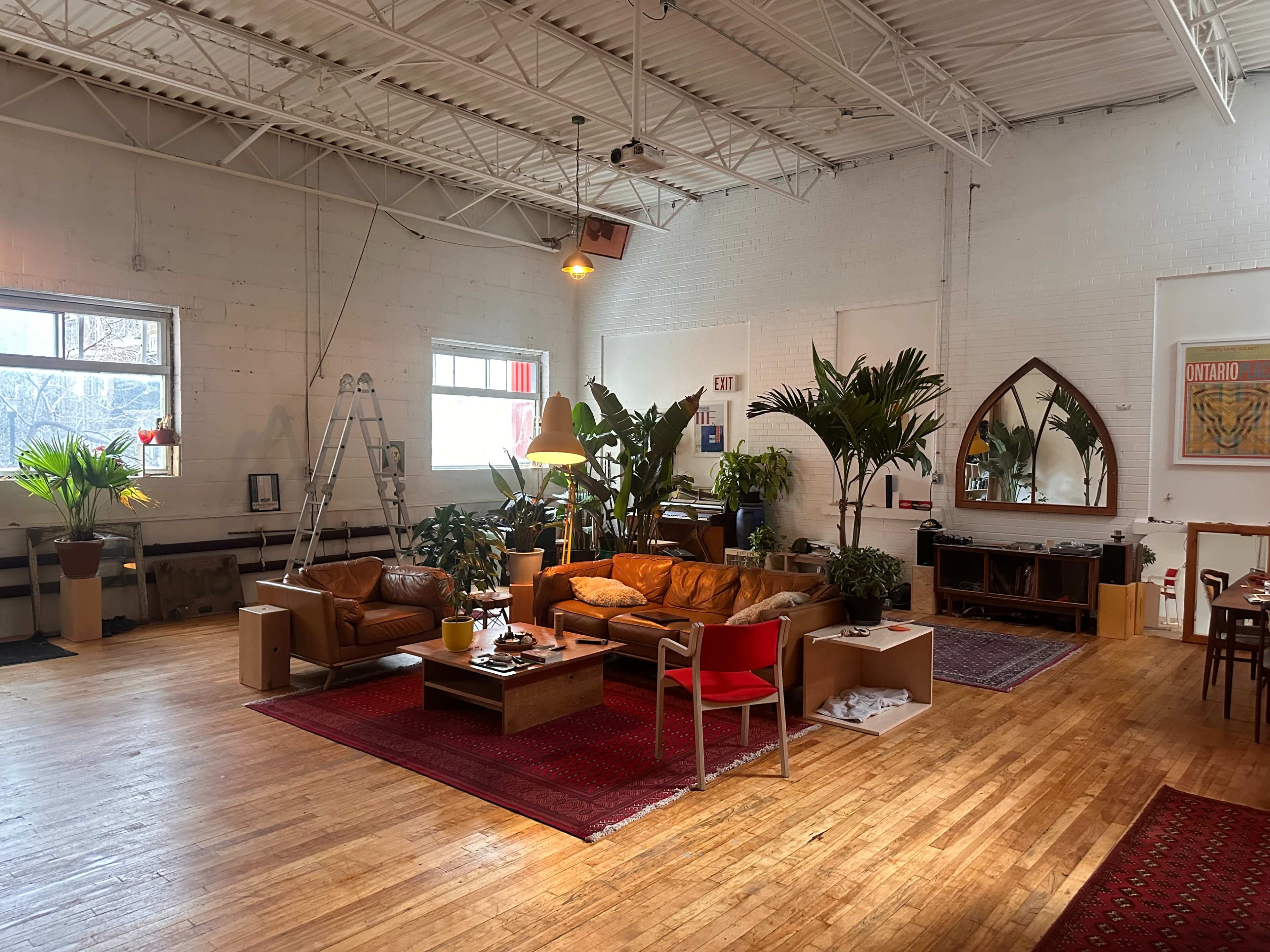 The image shows a spacious room with high ceilings, featuring a mix of seating areas, plants, a ladder, and wooden floors.