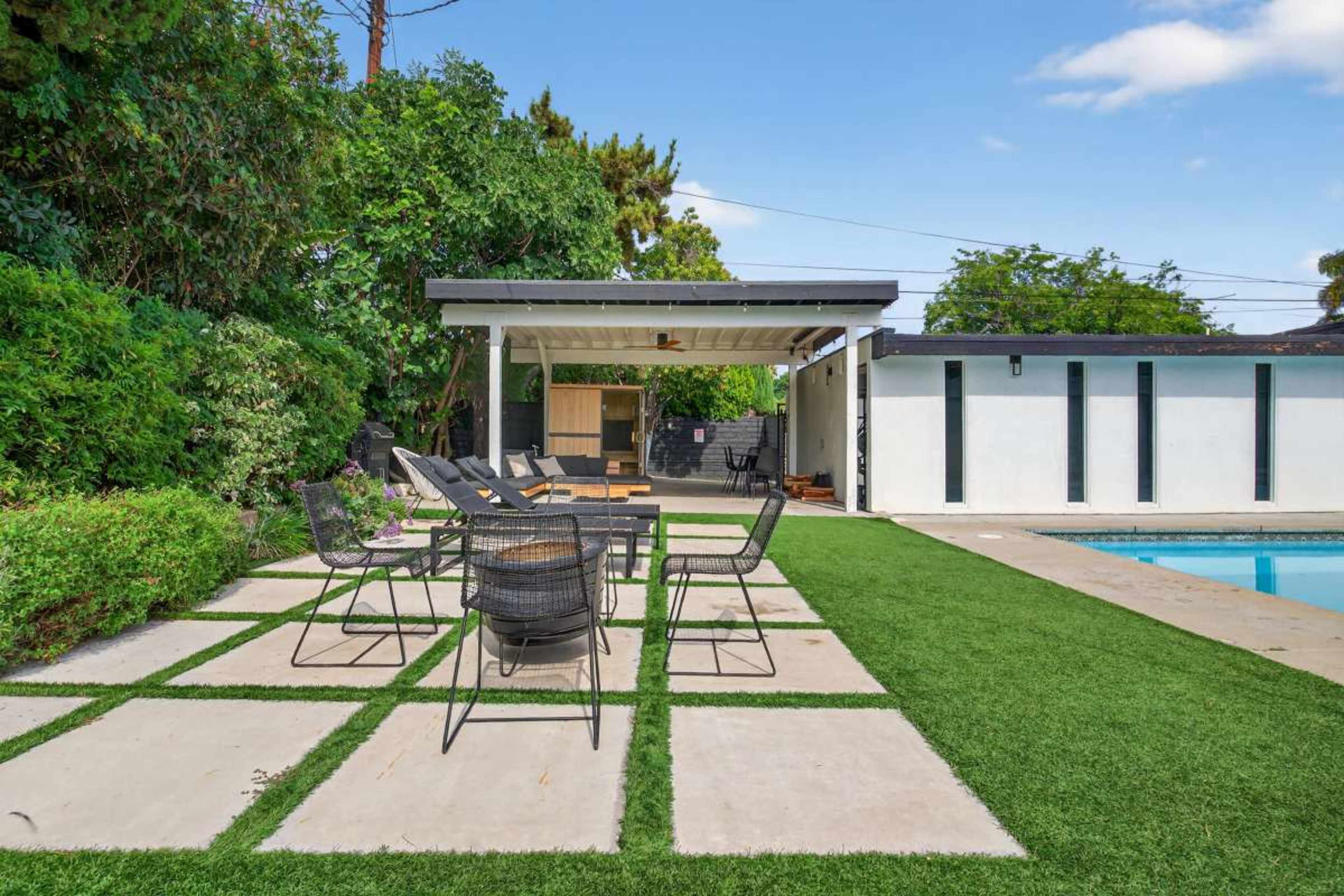 The image shows a backyard with a swimming pool, a patio area featuring a table and chairs, and a covered outdoor kitchen surrounded by greenery.
