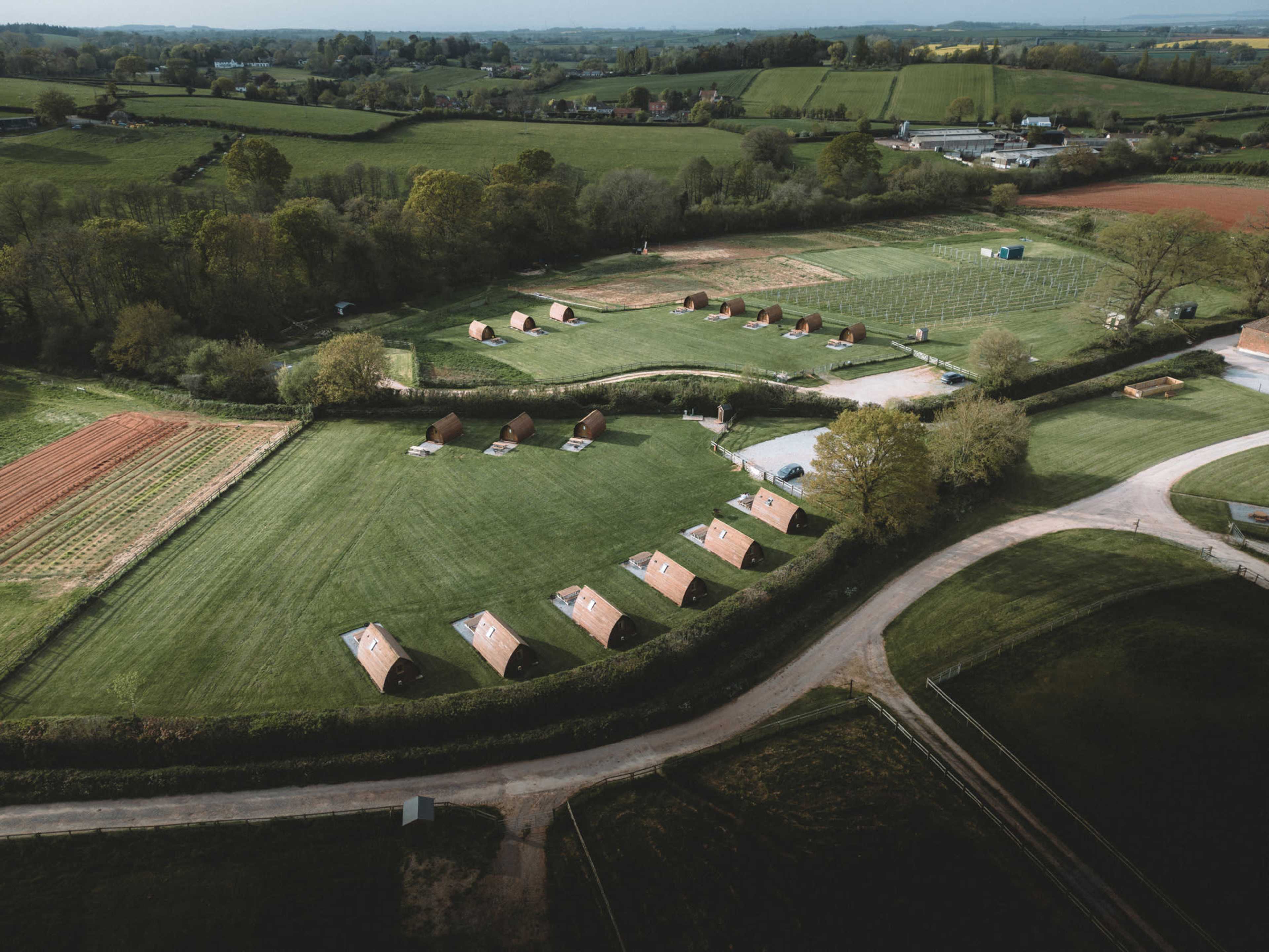 The image shows an aerial view of a countryside area with several tents arranged in a grassy field, surrounded by farmland and trees.