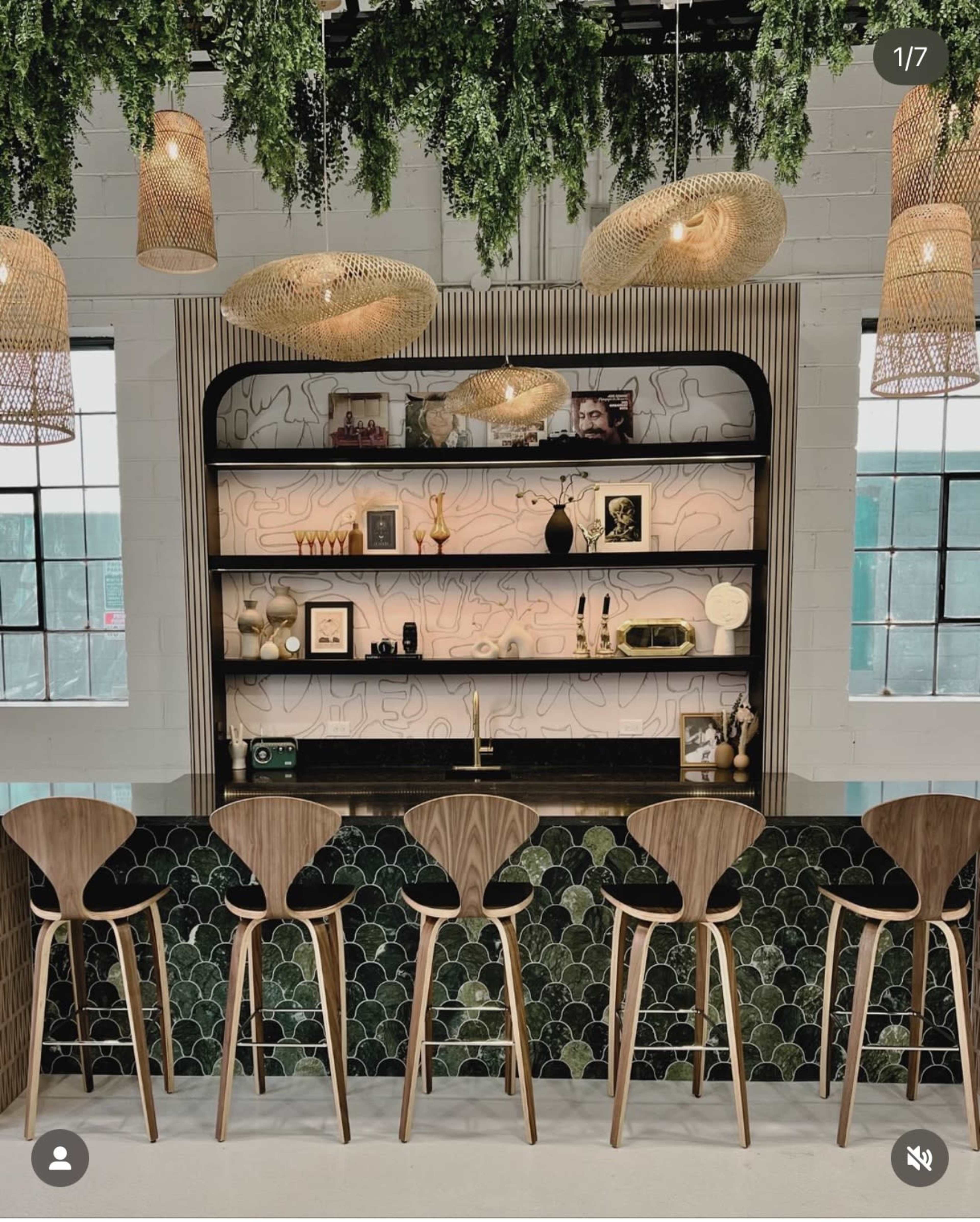 The image shows a modern bar area featuring wooden stools, a decorative backlit shelf with various items, and overhead lighting surrounded by greenery.