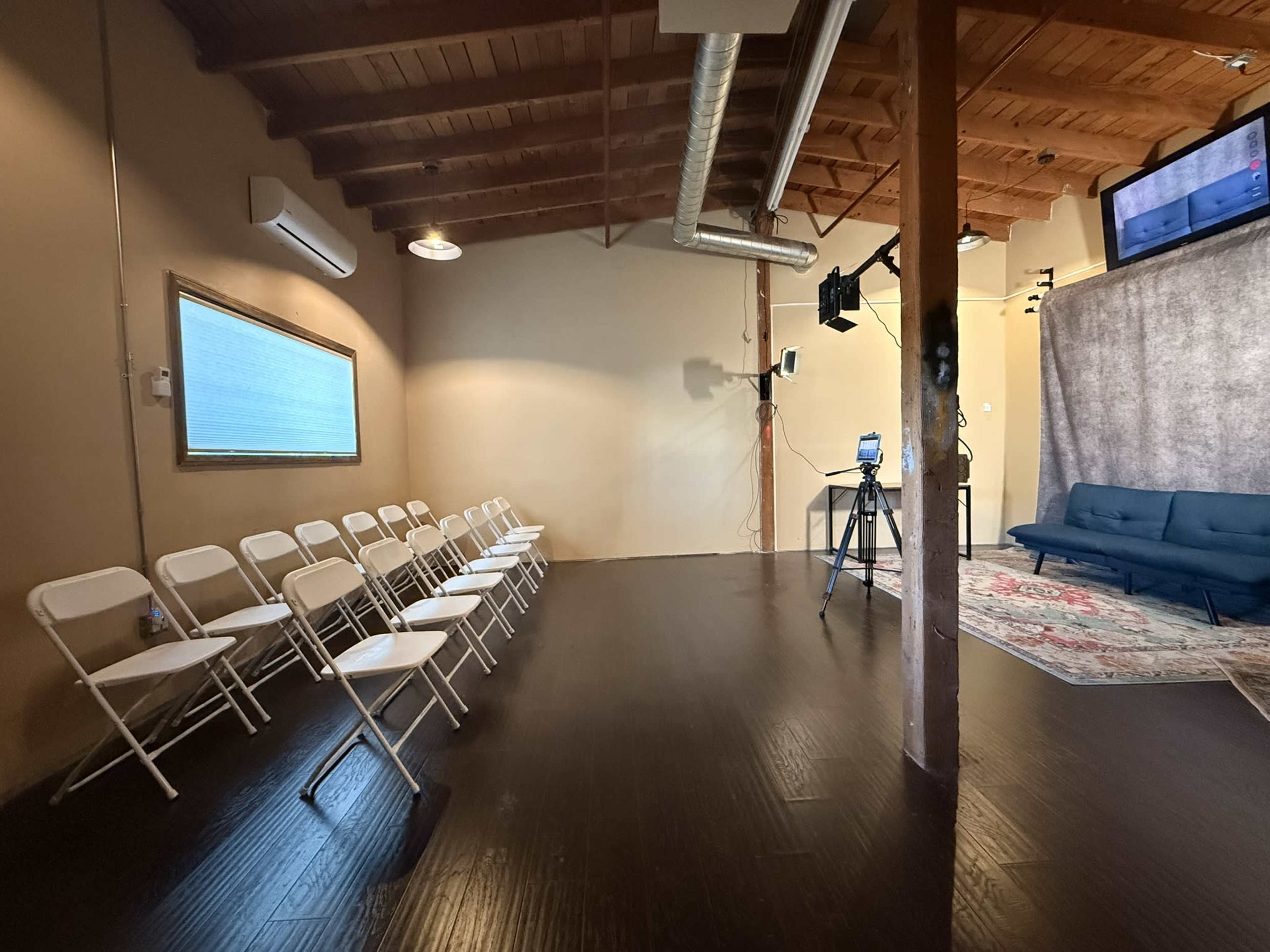 The image shows a sparsely furnished room featuring a row of white folding chairs, a blue couch, and a backdrop with a camera set up for a possible recording or presentation.