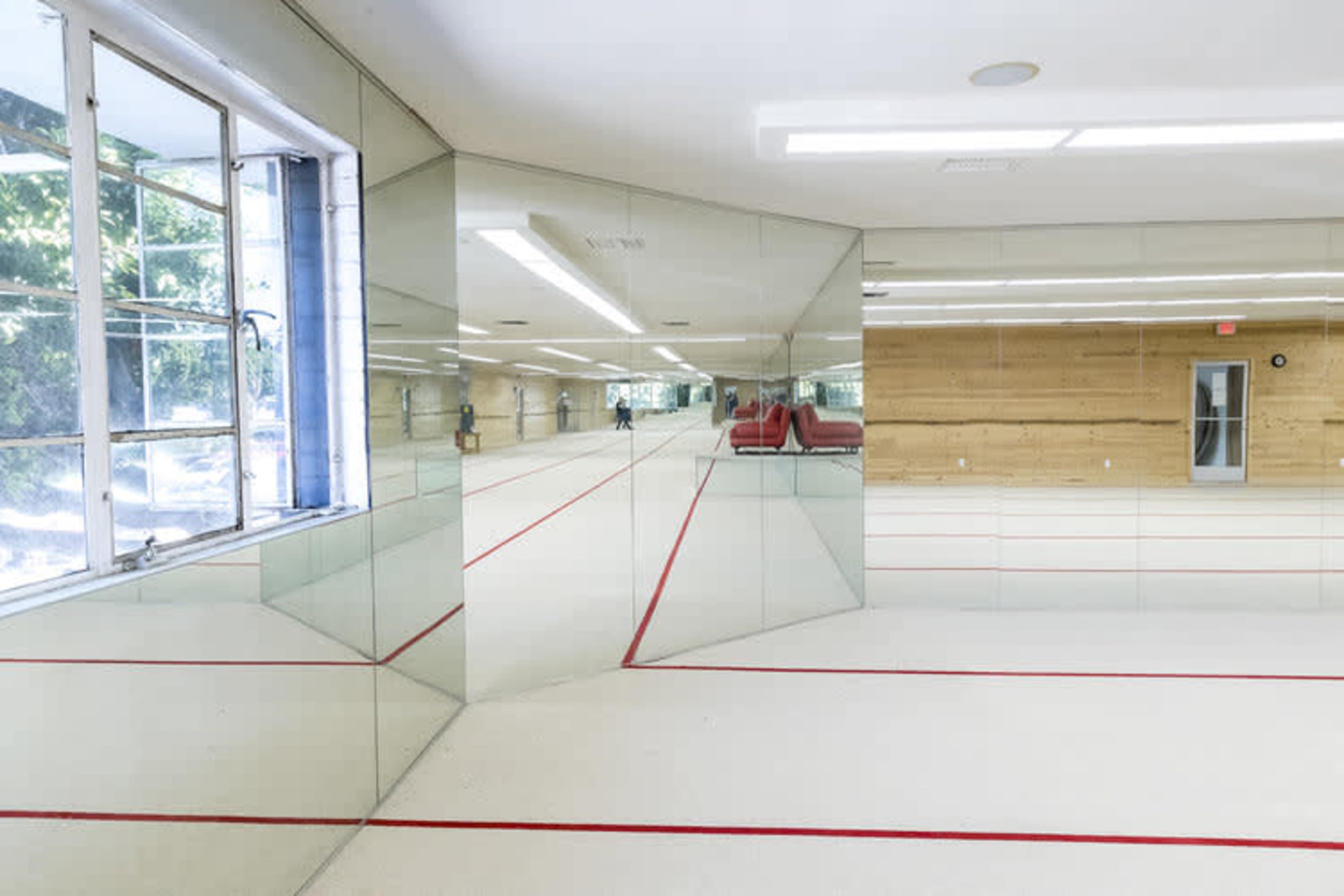 The image shows a brightly lit gymnasium with mirrored walls, a wooden accent wall, and a red-striped mat on the floor.