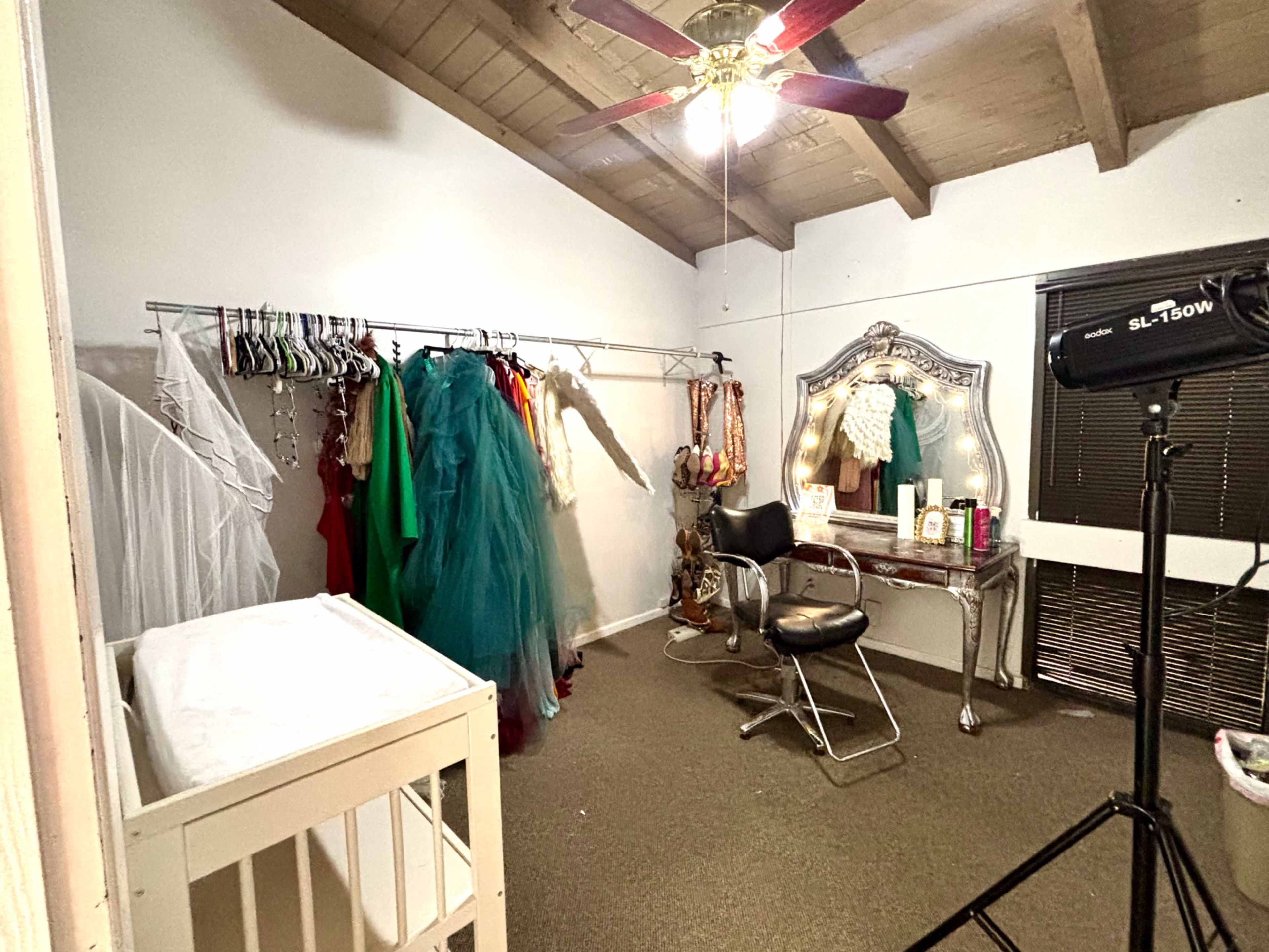 The image shows a room with a rack of colorful costumes, a mirrored vanity, a black chair, and a crib in one corner.