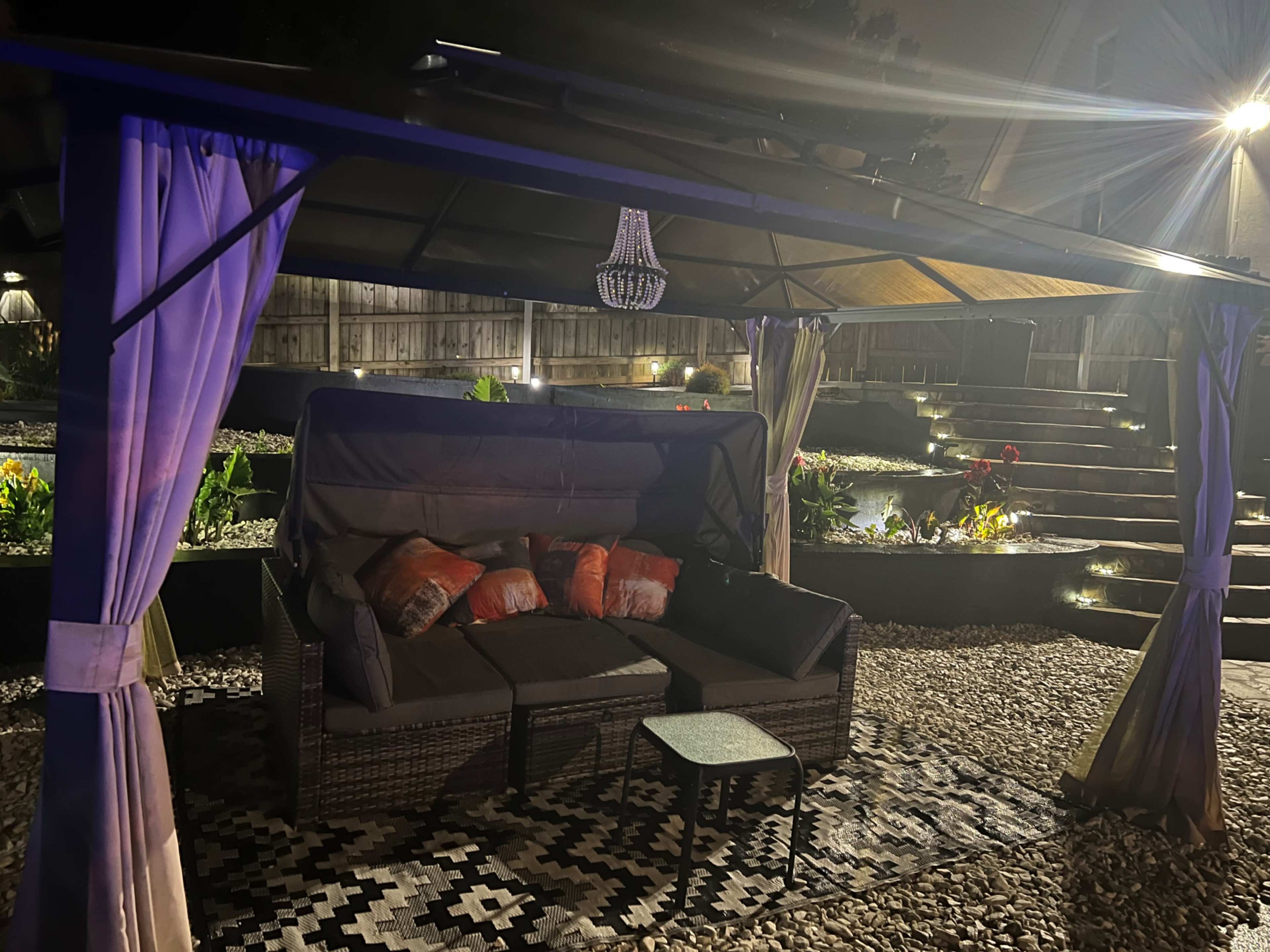 A dark outdoor setting features a gazebo with a seating area, adorned with cushions and a small table, surrounded by landscaped gardens and illuminated by soft lighting.