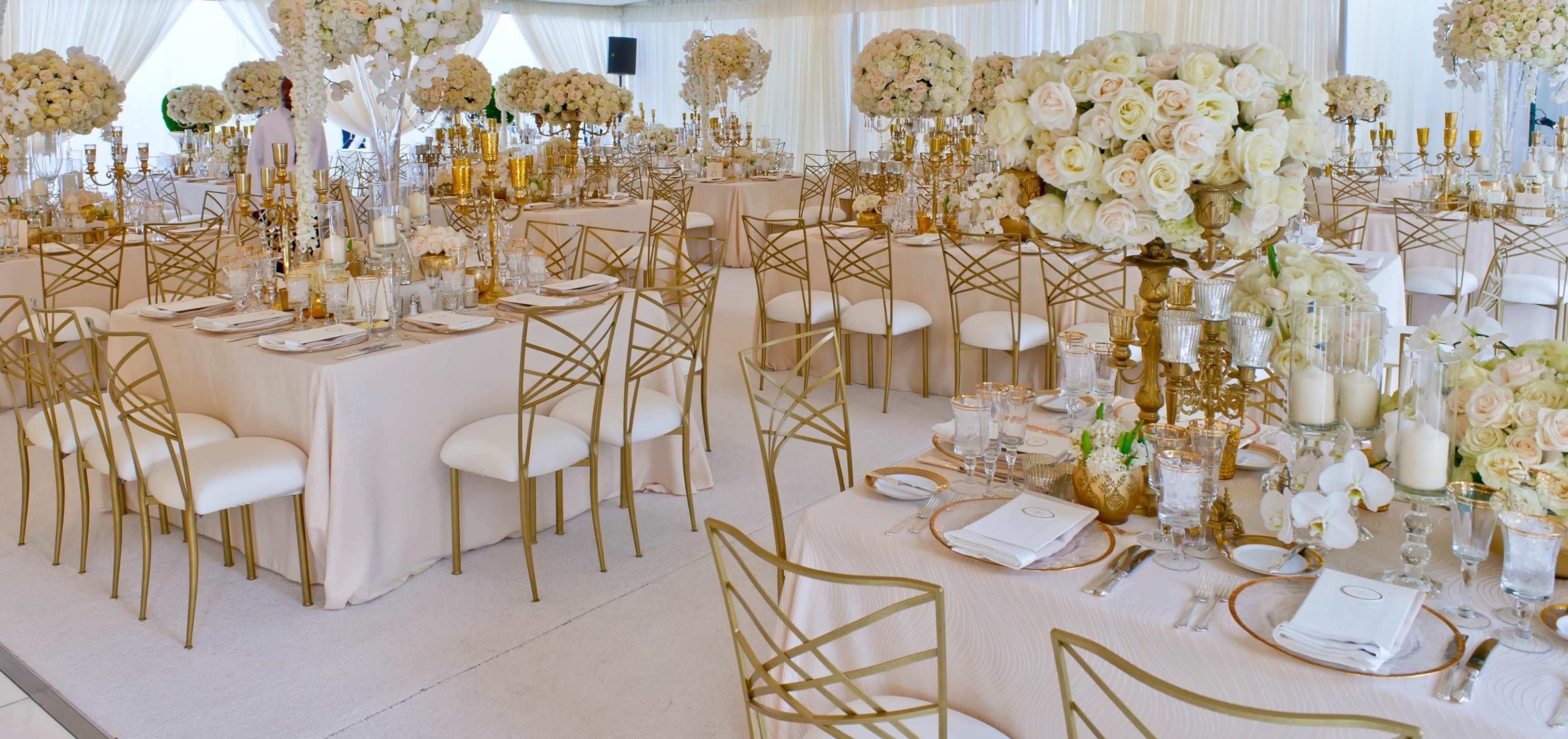 The image shows an elegantly arranged banquet hall with round tables set for a formal event, adorned with floral centerpieces and golden decorations.