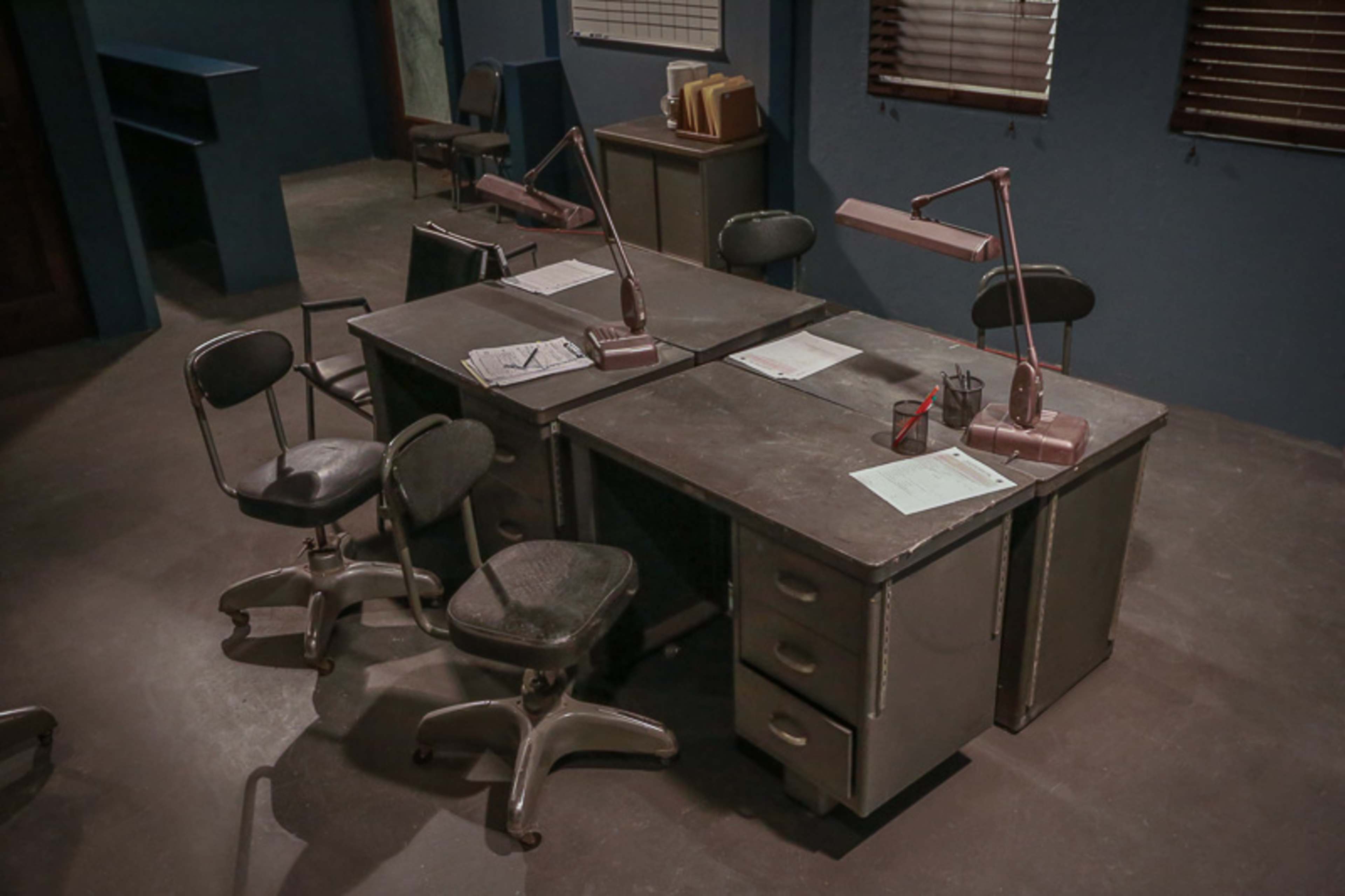 Two metal desks with attached lamps and office chairs in a room with blue walls and wooden blinds.