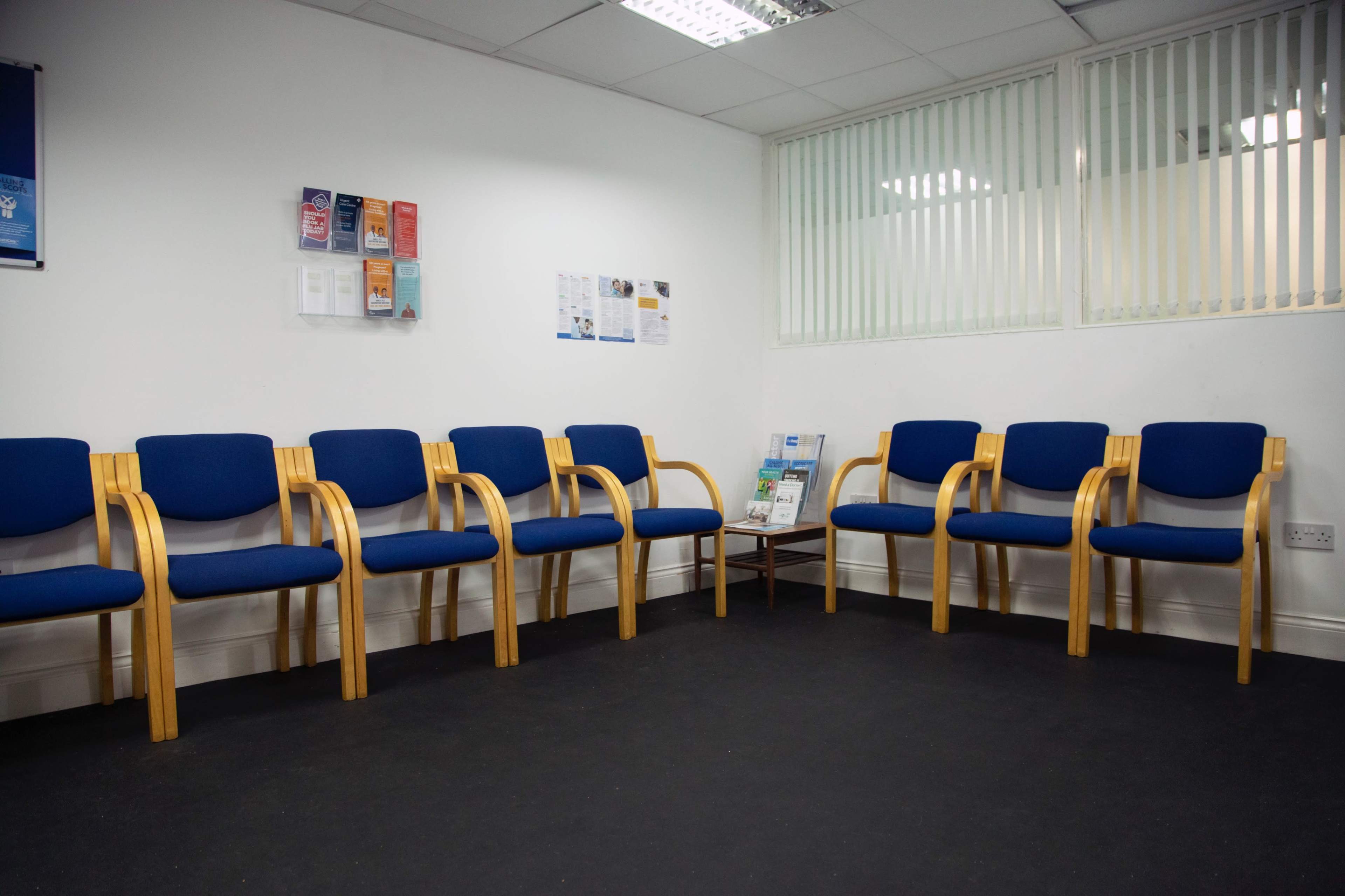 A waiting area with several blue chairs arranged in a semicircle against a white wall, with a small table holding brochures in the corner.