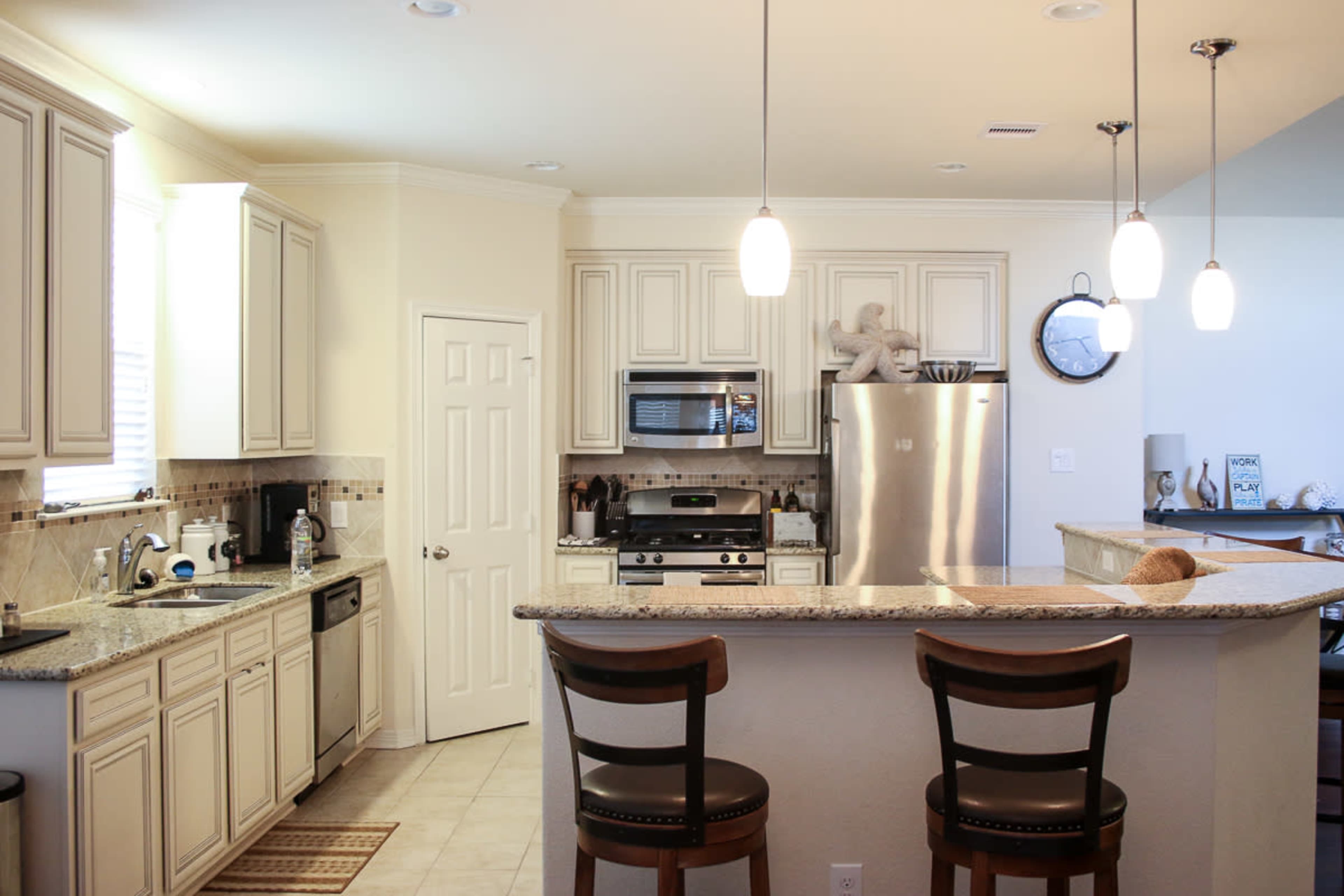 The image shows a modern kitchen with light-colored cabinetry, stainless steel appliances, and a granite countertop bar with two stools.