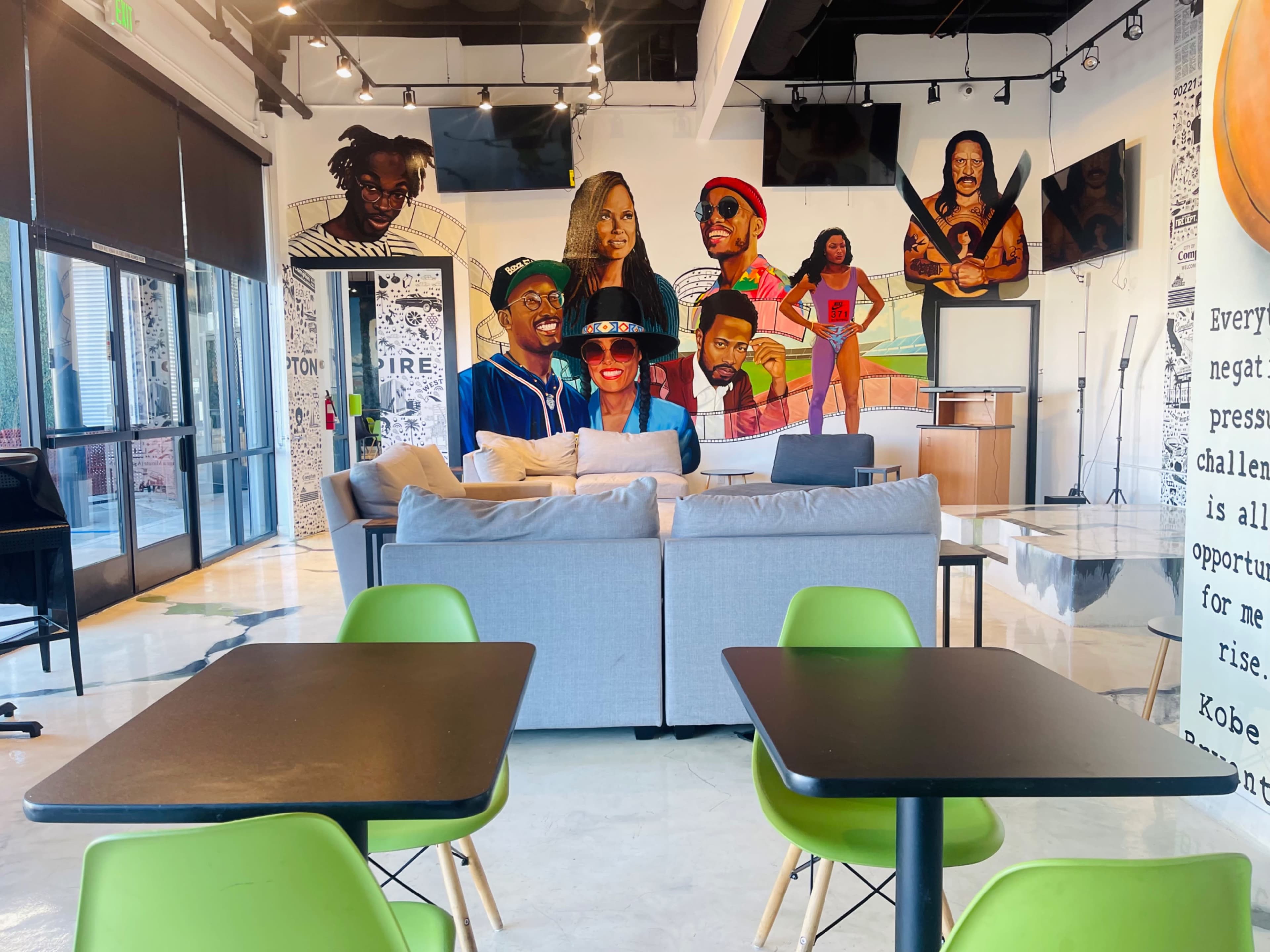 A modern cafe interior with a large mural featuring various characters on the wall behind a sofa and tables with green chairs.