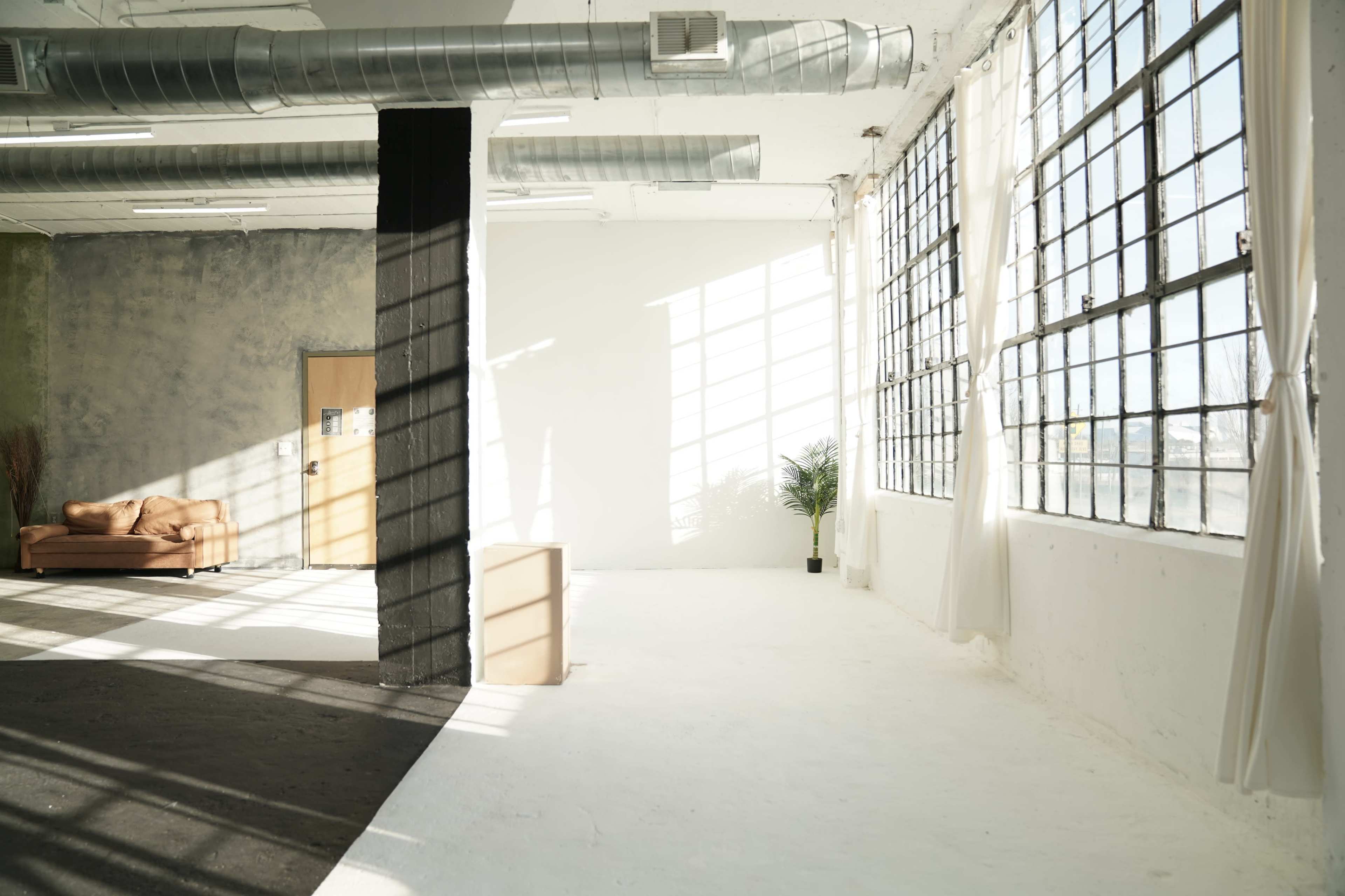 The image shows a bright, spacious room with large windows, a minimalist design, and a sofa in one corner.