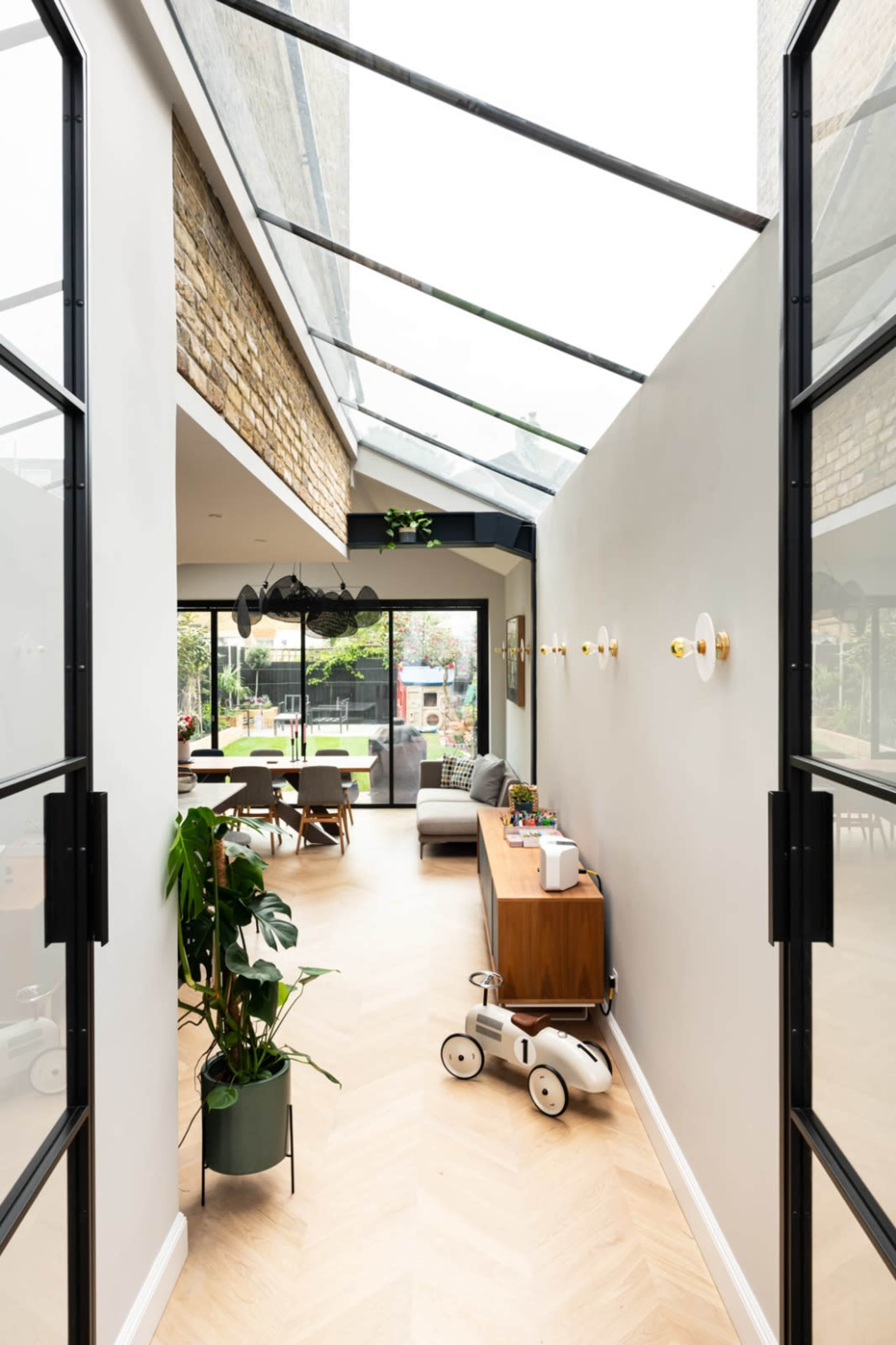 A well-lit hallway with glass walls leads to a spacious living area featuring modern furniture and greenery.