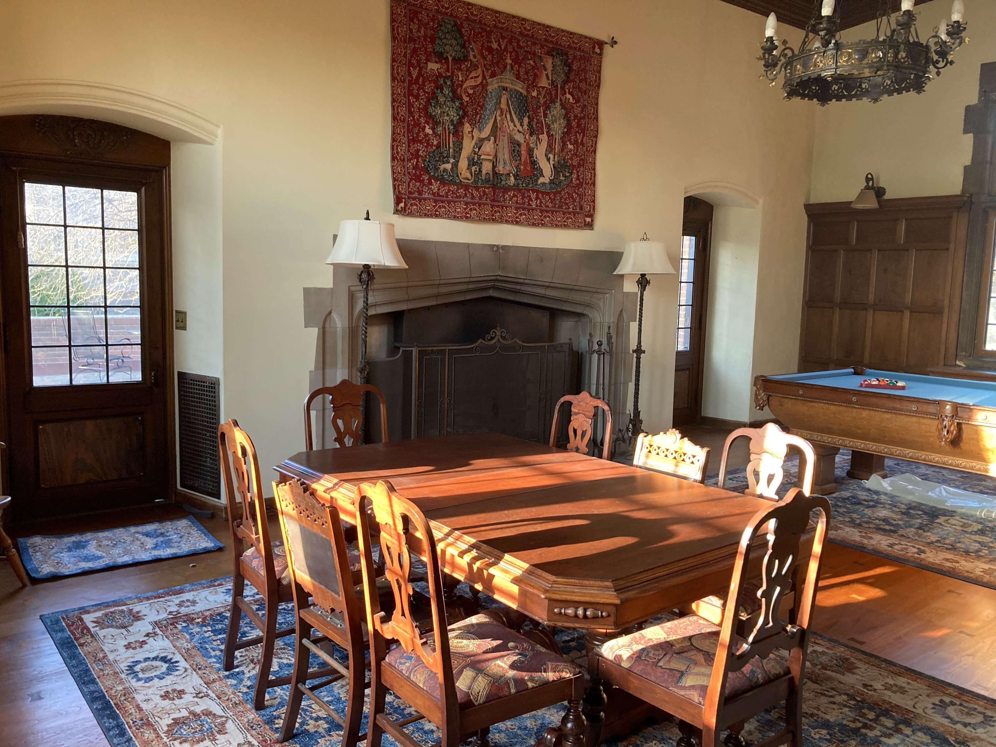 A well-furnished dining room with a wooden table surrounded by six chairs, a large tapestry hanging on the wall, and a pool table in the background.