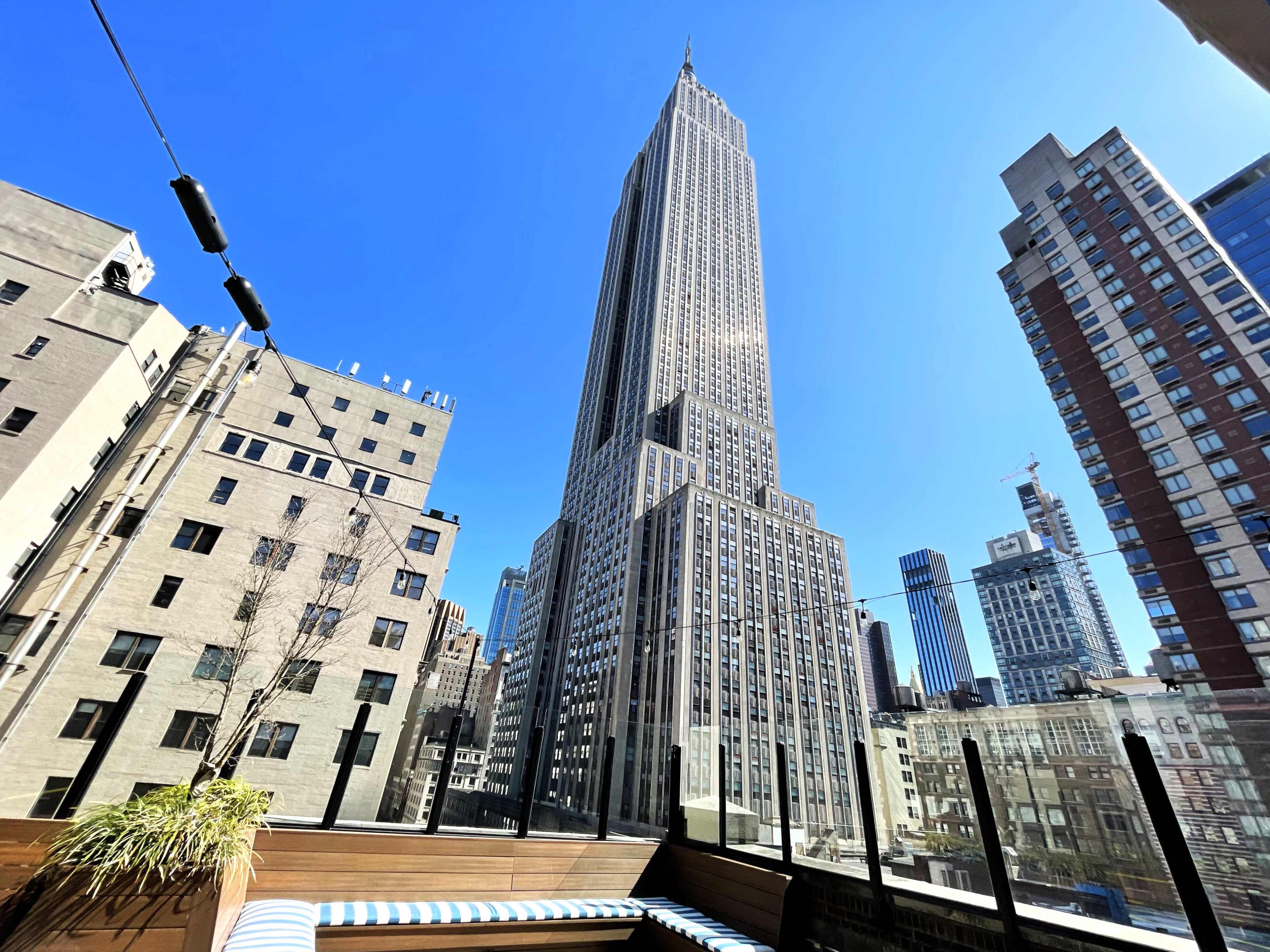 The image shows a view of the Empire State Building towering over a rooftop terrace in New York City under a clear blue sky.