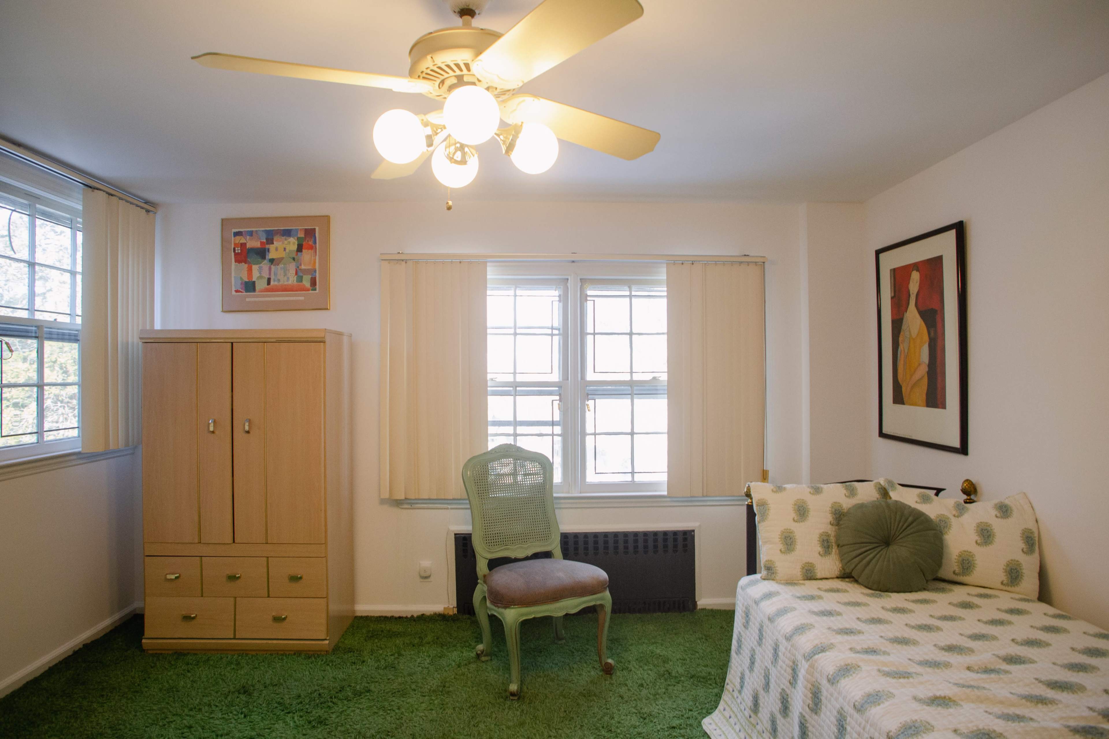 The room features a ceiling fan, a light green carpet, a daybed with patterned bedding, a wooden wardrobe, a decorative chair, and two windows with blinds.