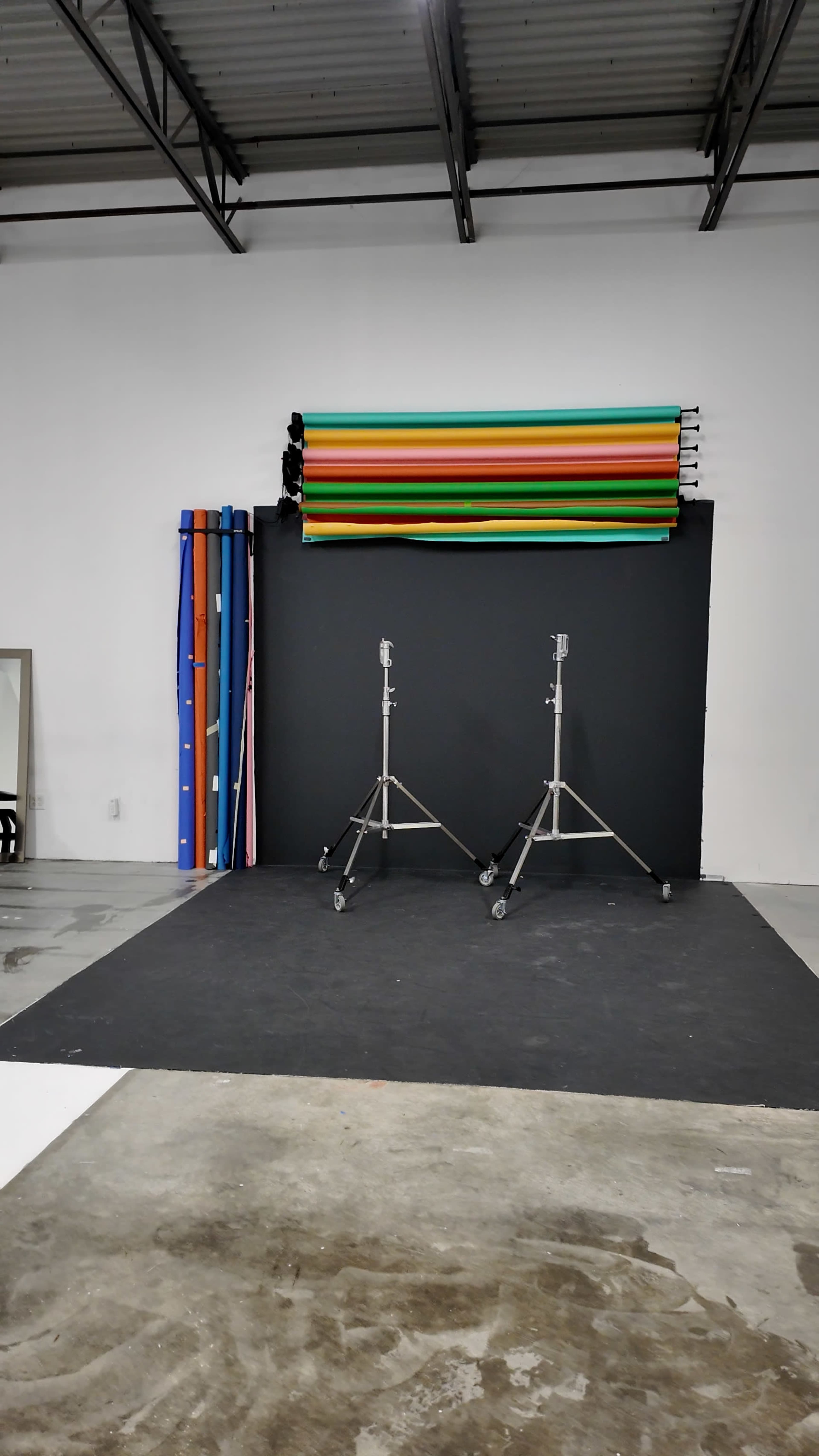 The image shows a photography studio setup with two lighting stands, a black backdrop, and colorful backdrop rolls stacked on a wall.