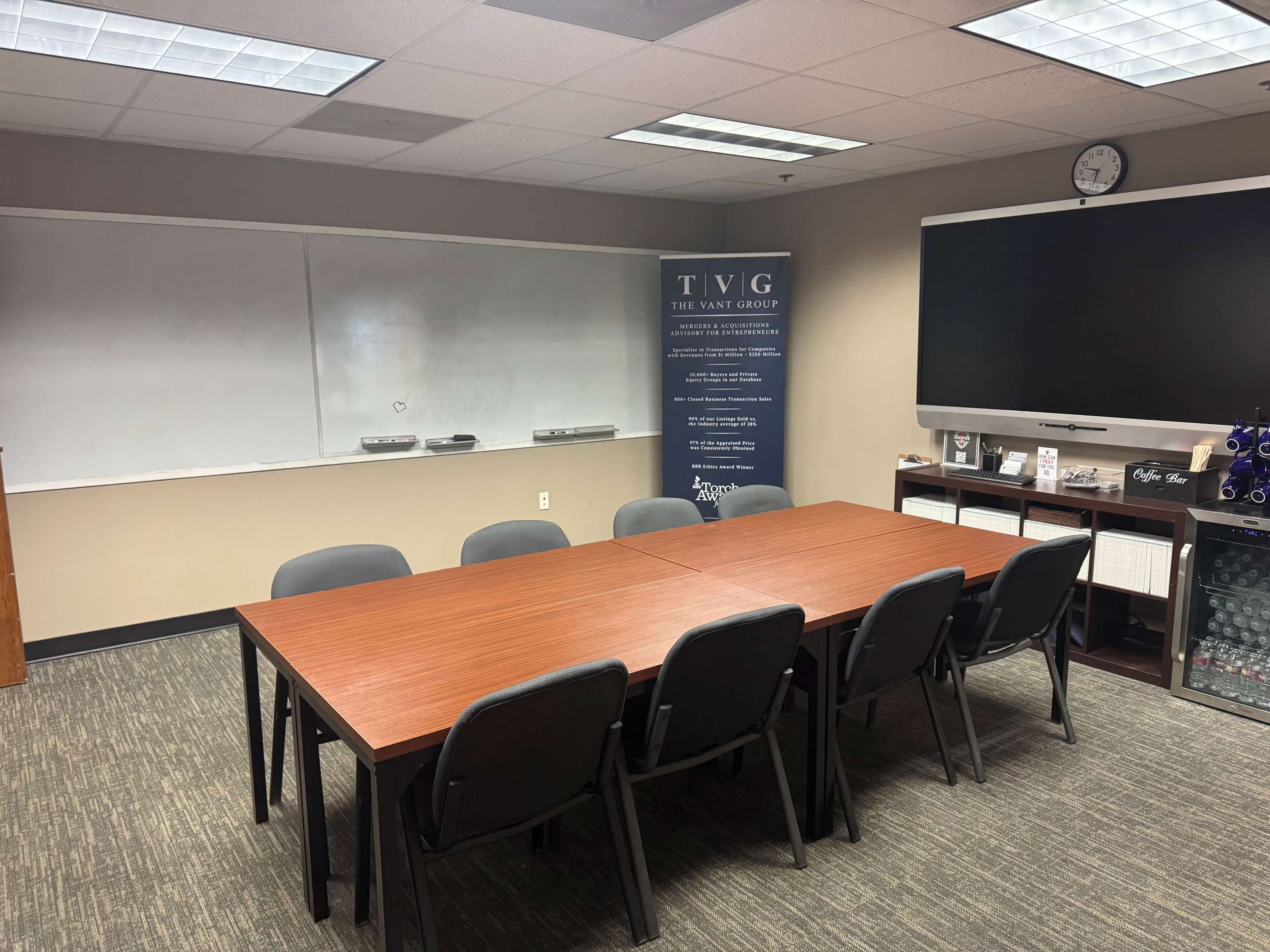 A conference room features a long wooden table surrounded by six chairs, a large television screen, and a whiteboard on one wall.