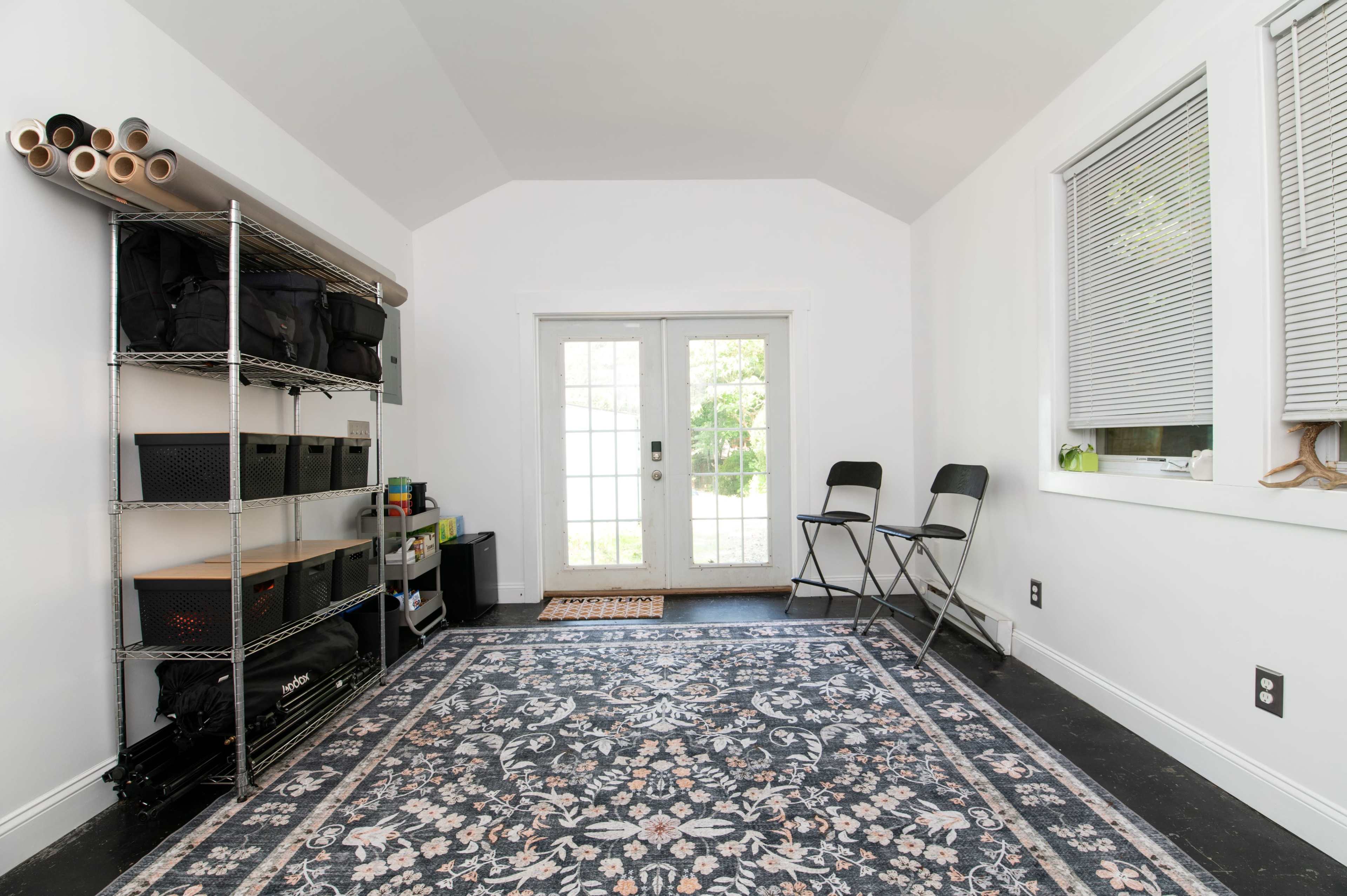 The image shows a neatly organized room with a patterned area rug, a metal shelving unit, two folding chairs, and large front doors leading outside.