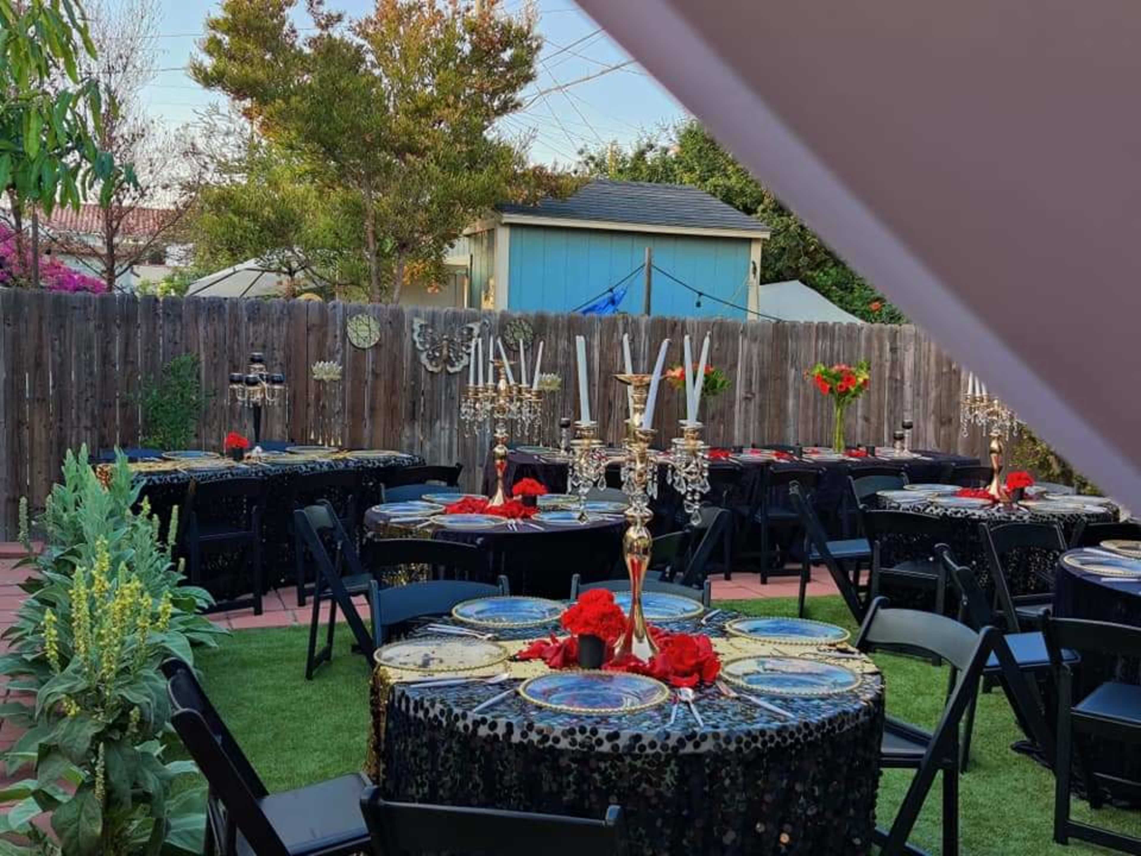 A backyard is set up for a party with several elegantly decorated tables featuring black tablecloths, floral centerpieces, and candelabras.