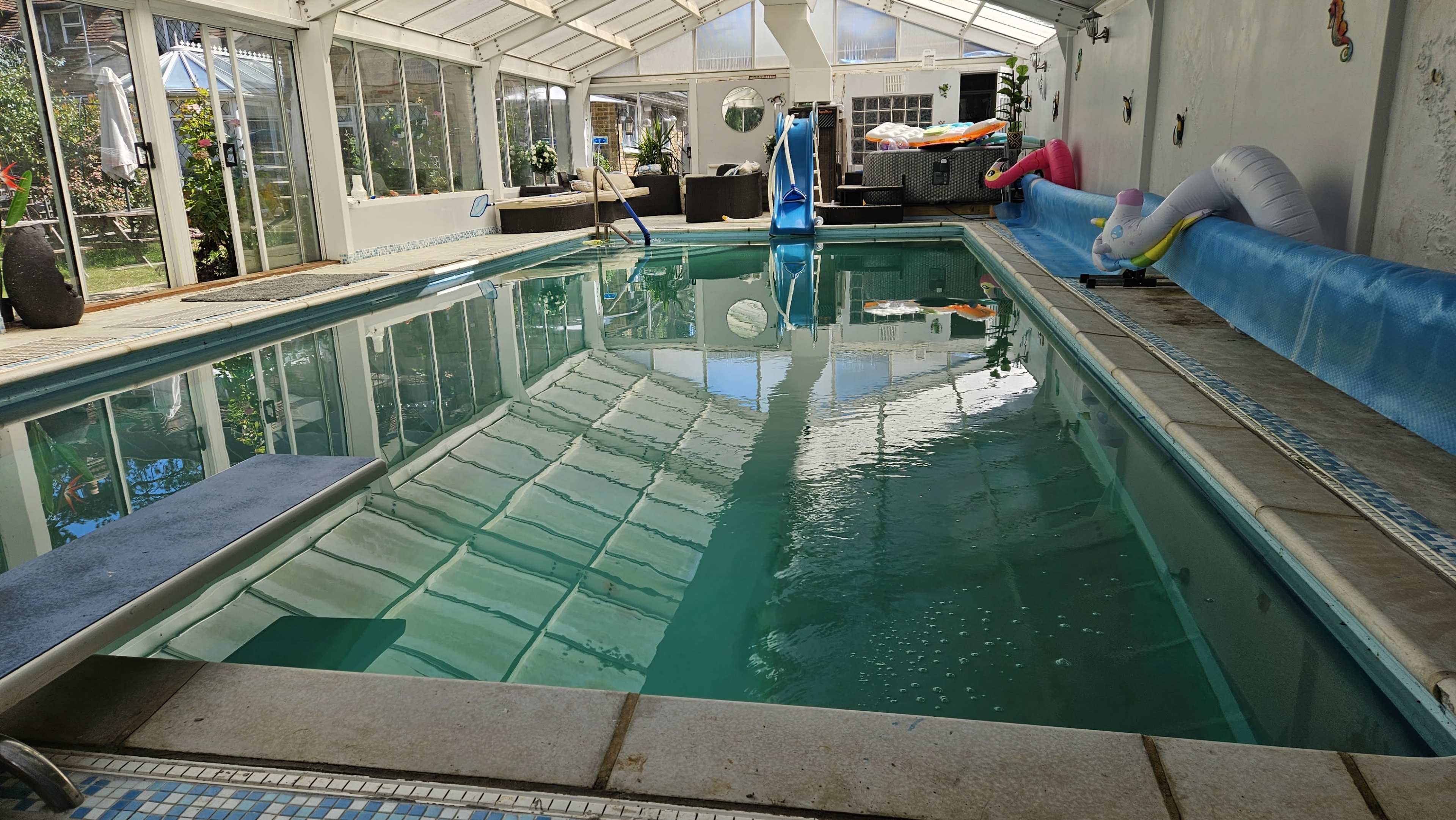 The image shows an indoor swimming pool with clear water, surrounded by a glass enclosure and various colorful pool toys.