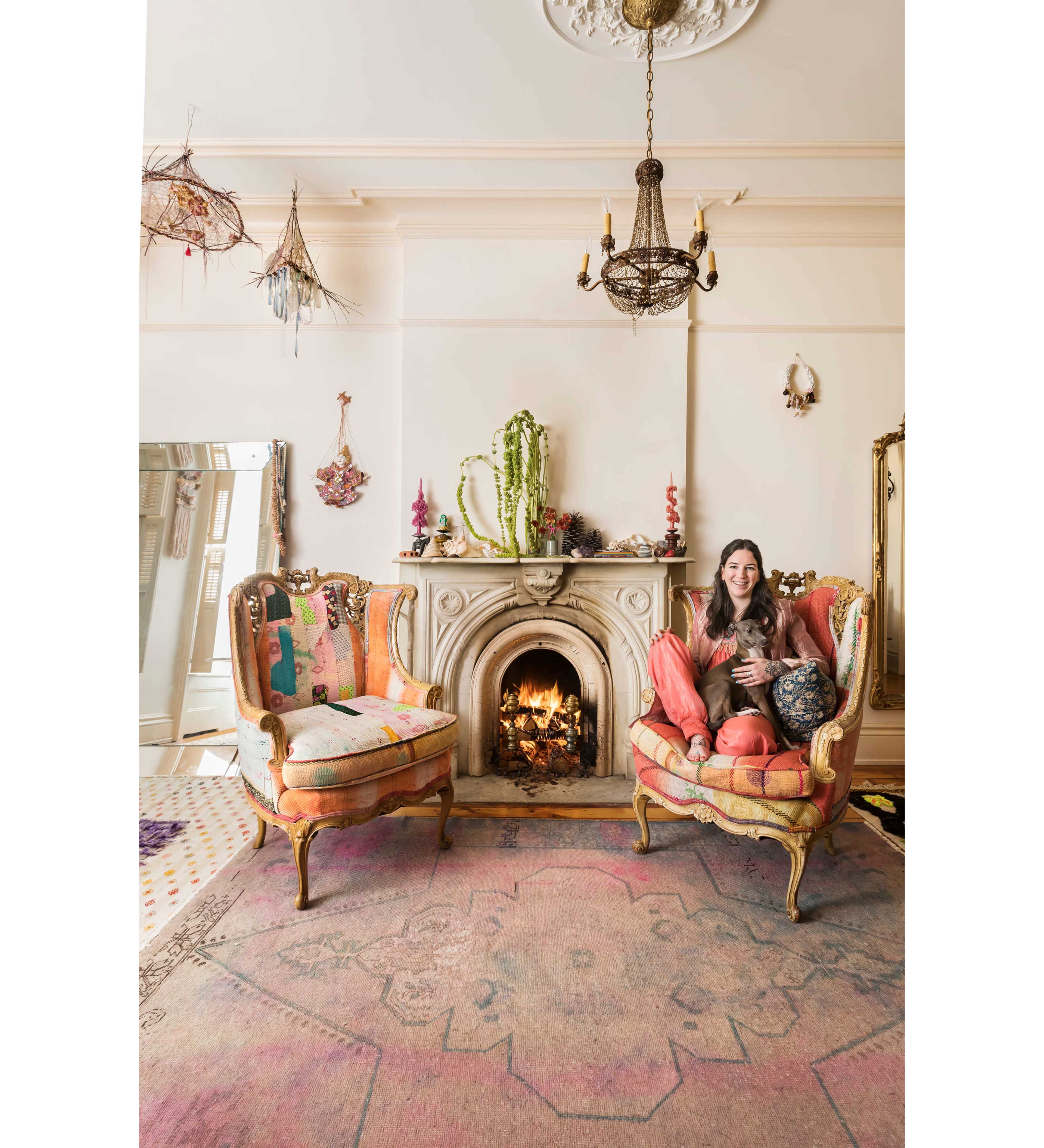 A woman sits comfortably in a colorful armchair beside a decorative fireplace, surrounded by eclectic home decor and a large mirror.