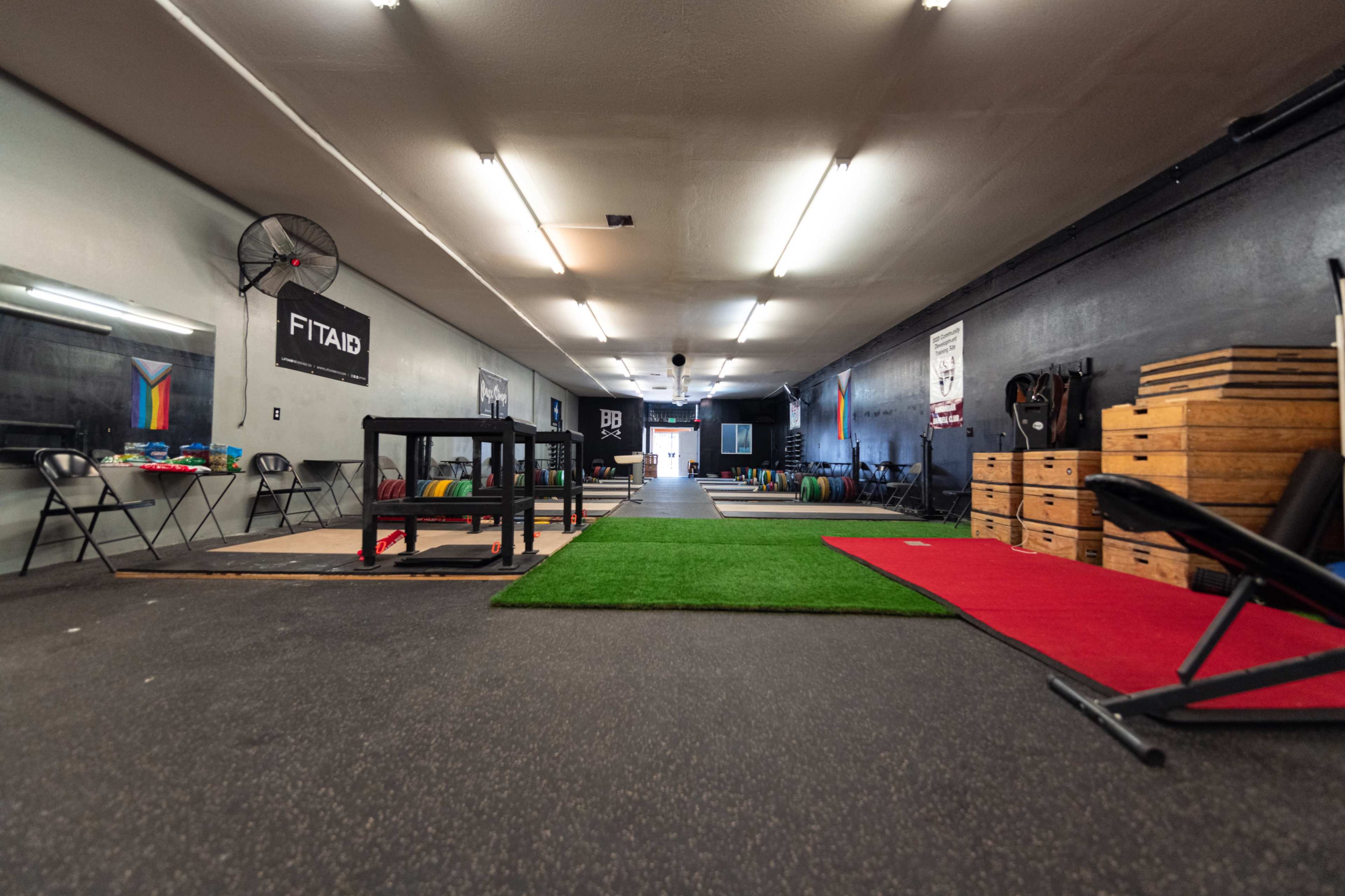 The interior of a fitness gym with a long hallway, exercise equipment on the sides, and various flooring surfaces including artificial grass and mats.