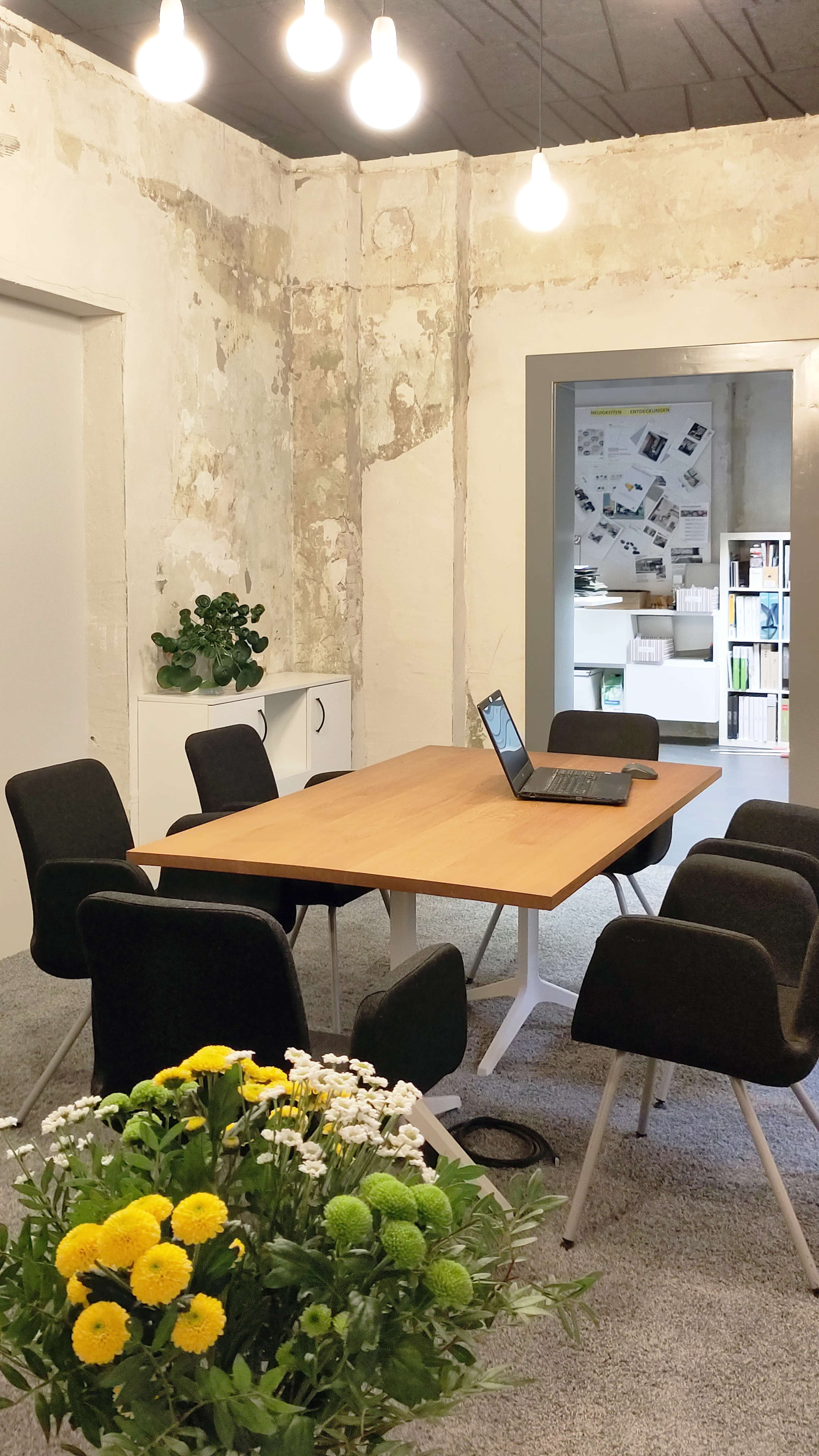 A wooden table with a laptop is surrounded by six chairs in a room with exposed walls and a view of a neighboring area filled with office materials.
