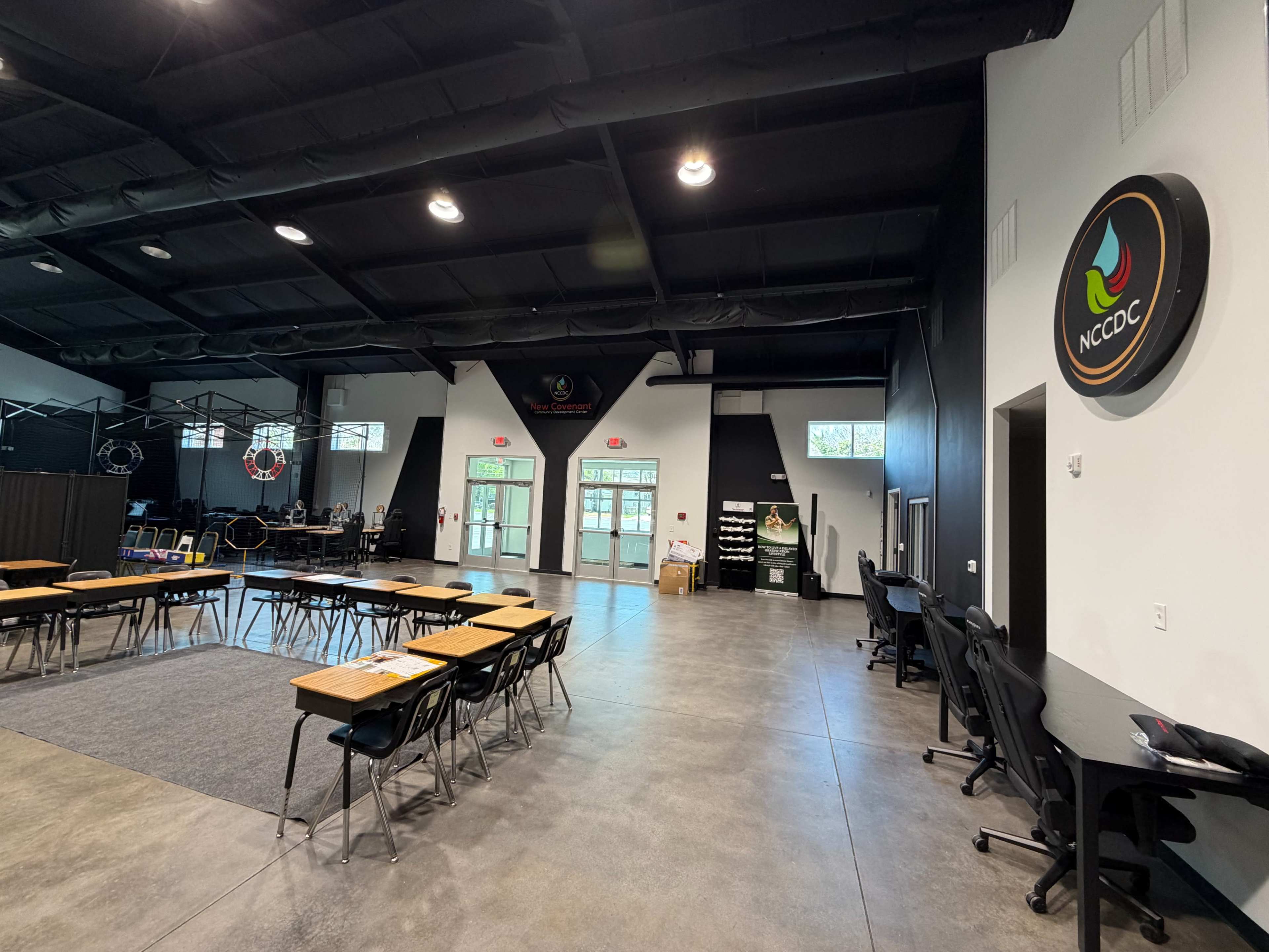 The image shows a spacious interior of a modern facility with tables, chairs, and a logo for NCCDC on the wall.