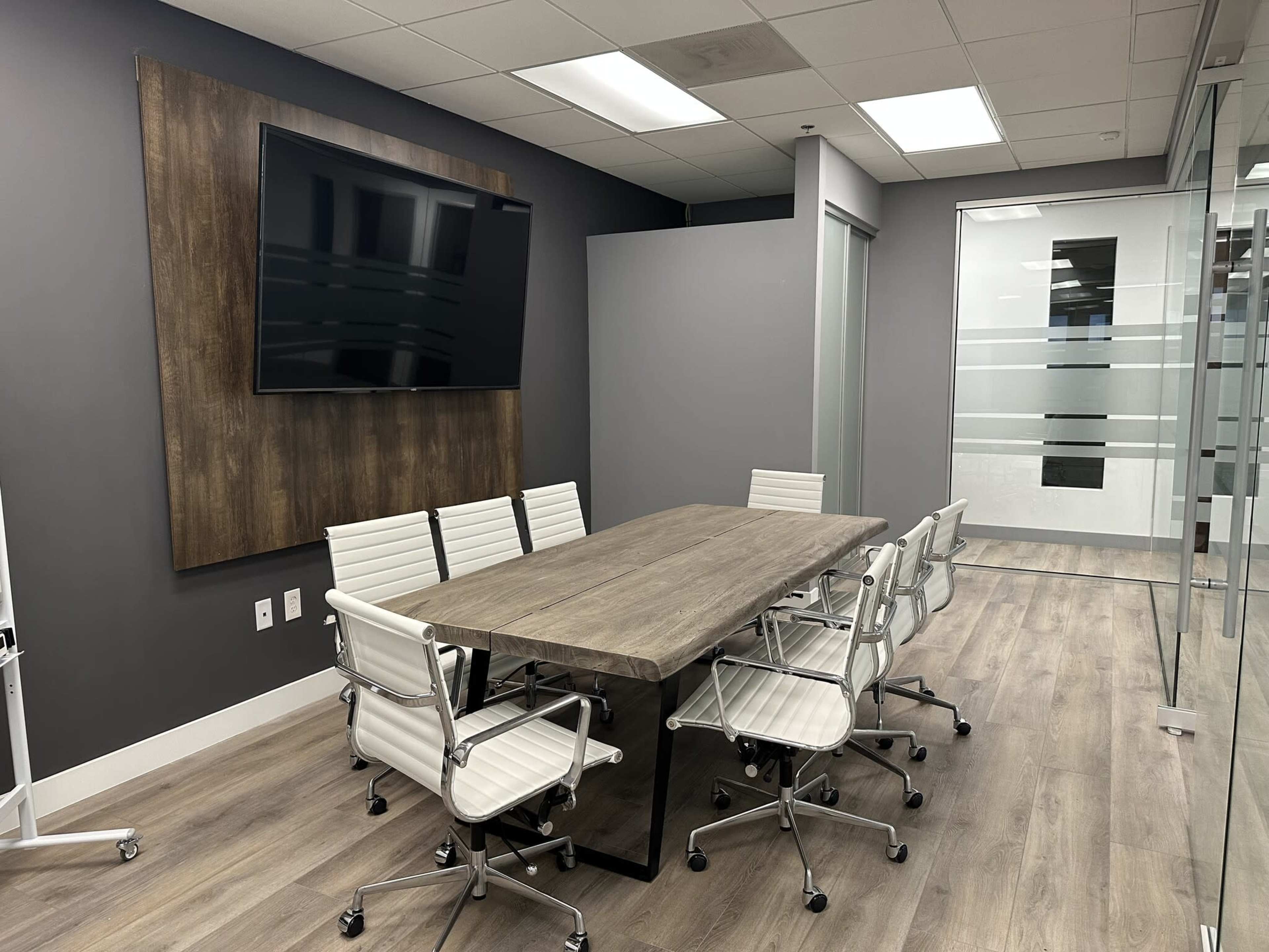 A modern conference room features a large wooden table surrounded by white rolling chairs, with a wall-mounted TV and glass partitions.