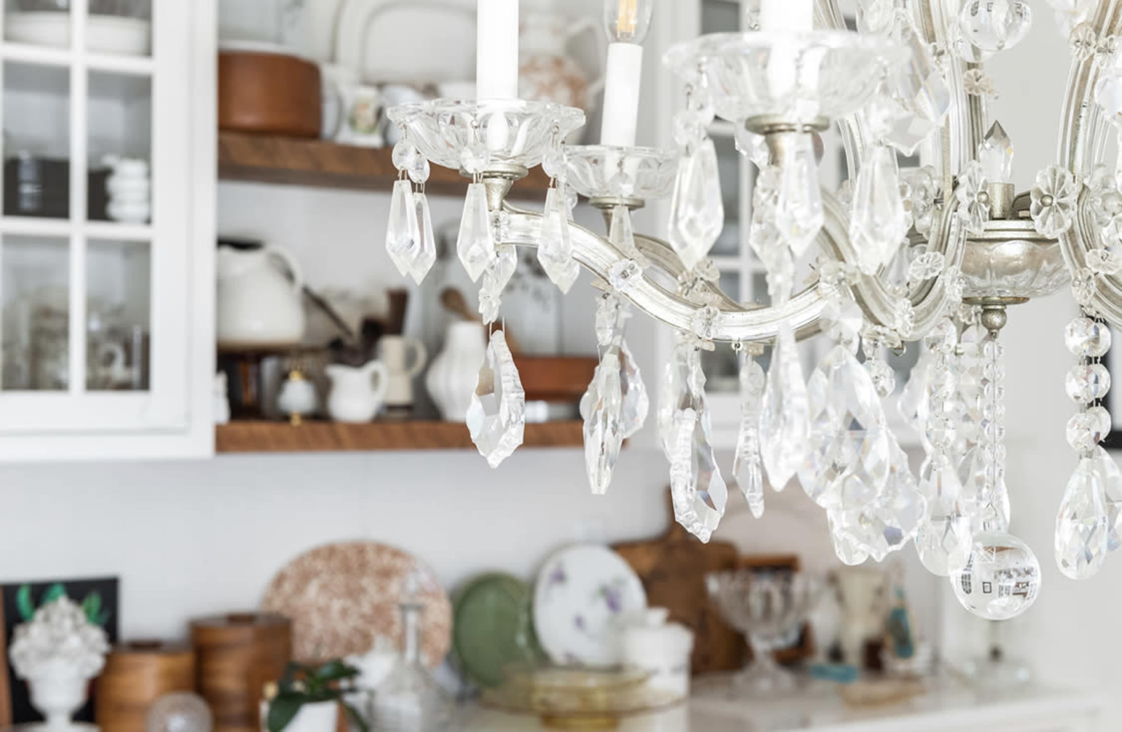 A crystal chandelier hangs above a shelf filled with various dishes and decorative items in a brightly lit kitchen.