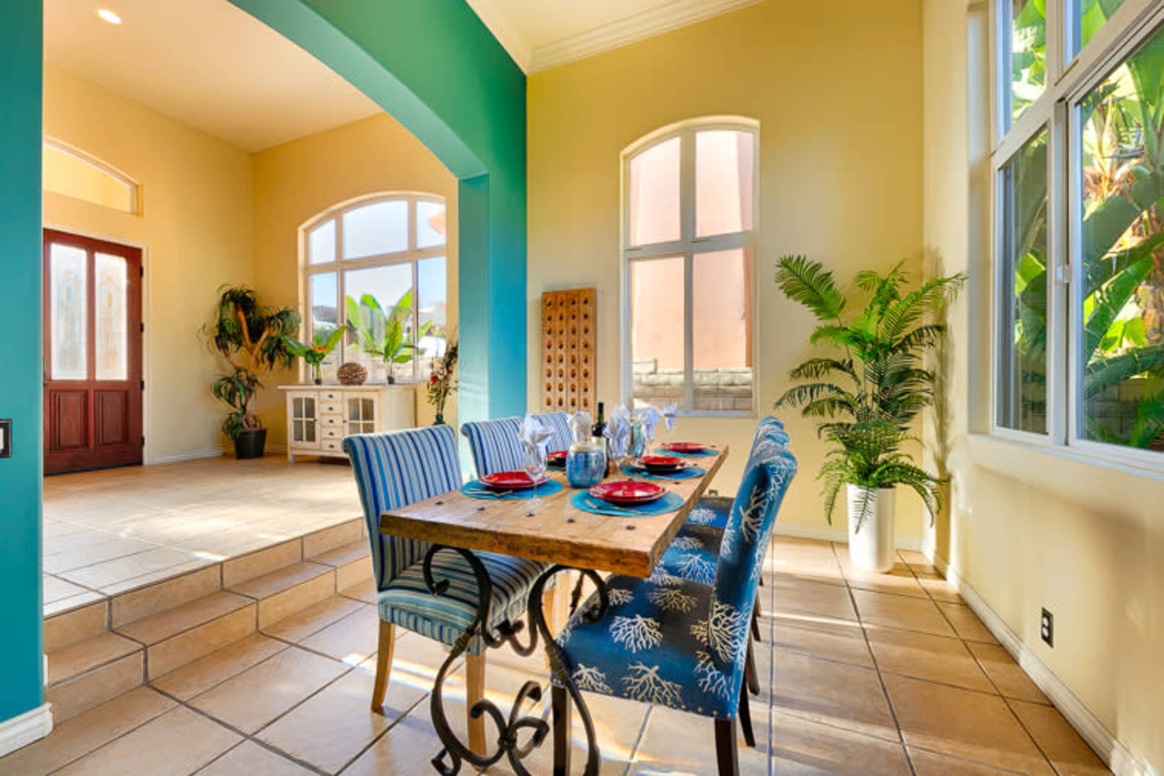 A dining area features a wooden table set with plates and glasses, surrounded by blue and white patterned chairs, beside large windows and greenery.